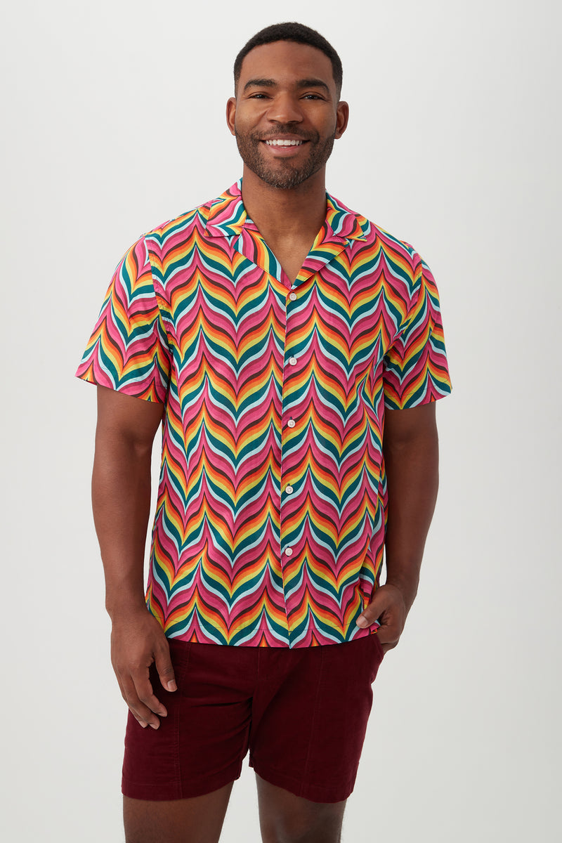 A man smiles while wearing the Felix Shirt in Multi, a brightly colored short-sleeve camp shirt with a bold rainbow zigzag pattern and revere collar, paired with dark red shorts, standing against a plain light background.