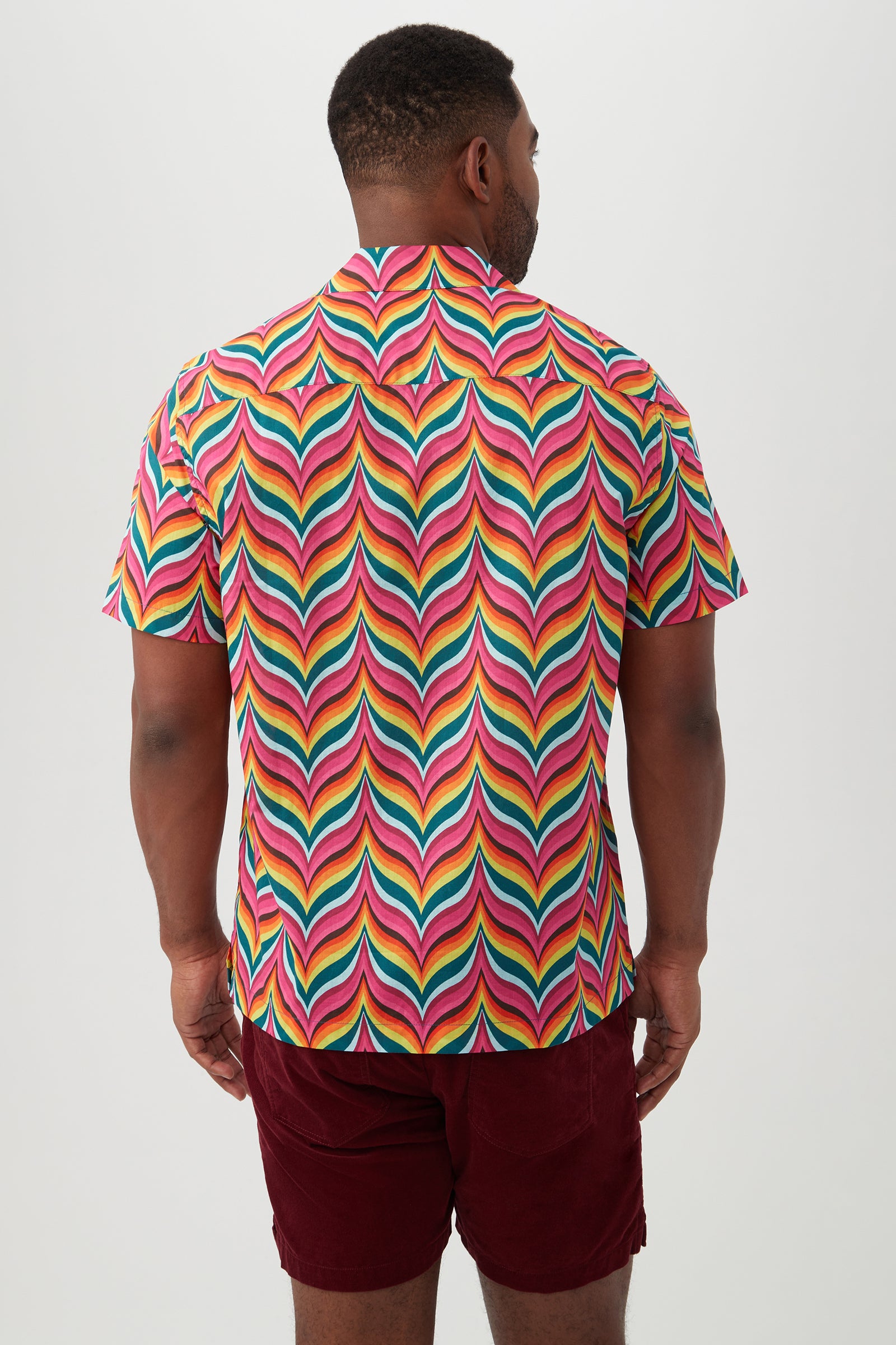 A man stands facing away, wearing the Felix Shirt—a colorful camp shirt with a revere collar and vibrant, wavy pink, yellow, green, and blue pattern—paired with burgundy shorts against a plain light background.
