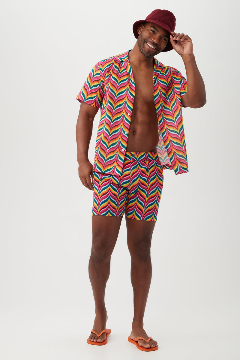 A smiling man in a red hat wears the Felix Shirt—a colorful, zigzag-patterned camp shirt with a revere collar and matching organic cotton shorts—paired with orange flip-flops, standing against a plain white background.