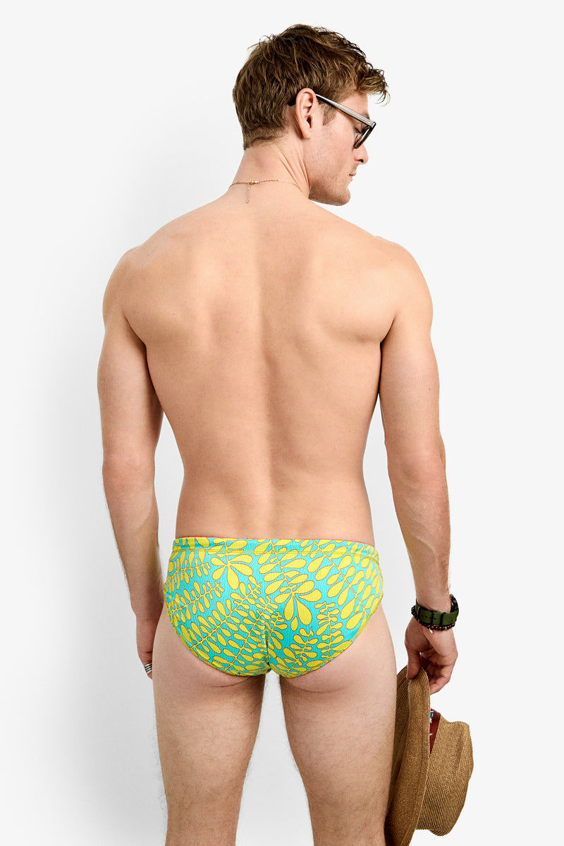 A man wearing glasses and the PALM BEACH SWIM BRIEF, a green-and-blue patterned reversible swim brief, stands facing away holding a hat. He is shirtless, wears a wristwatch, and displays this stylish men's swimwear.