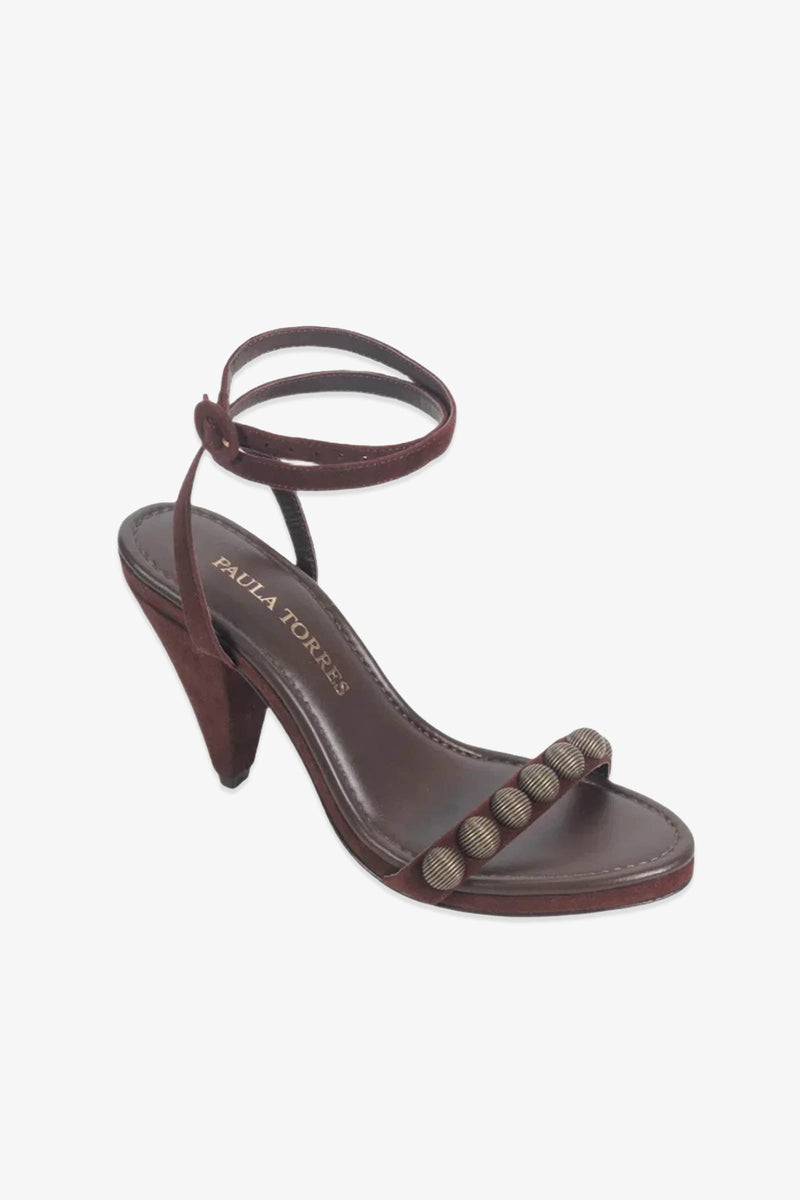 The Paula Torres Espanha Sandal is a brown high-heeled sandal with an ankle strap and five metallic beads adorning the toe strap. It features an architectural triangle heel, a cushioned insole, and showcases the signature PAULA TORRES label.