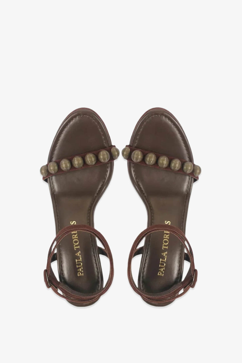 Introducing the Paula Torres Espanha Sandal: a brown open-toe design featuring a beaded strap across the toes and elegant ankle straps. The sandals are enhanced with an architectural triangle heel and a cushioned insole stamped with PAULA TORRES in gold, offering a perfect balance of style and comfort.