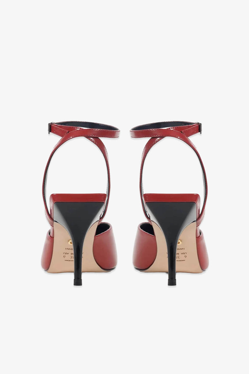 A pair of Paula Torres Cannes Pump high-heeled shoes with ankle straps, displayed from the back against a white background. With their elegant pointed toes, black heels and soles, and glossy red finish, these luxury pumps exude sophisticated appeal.