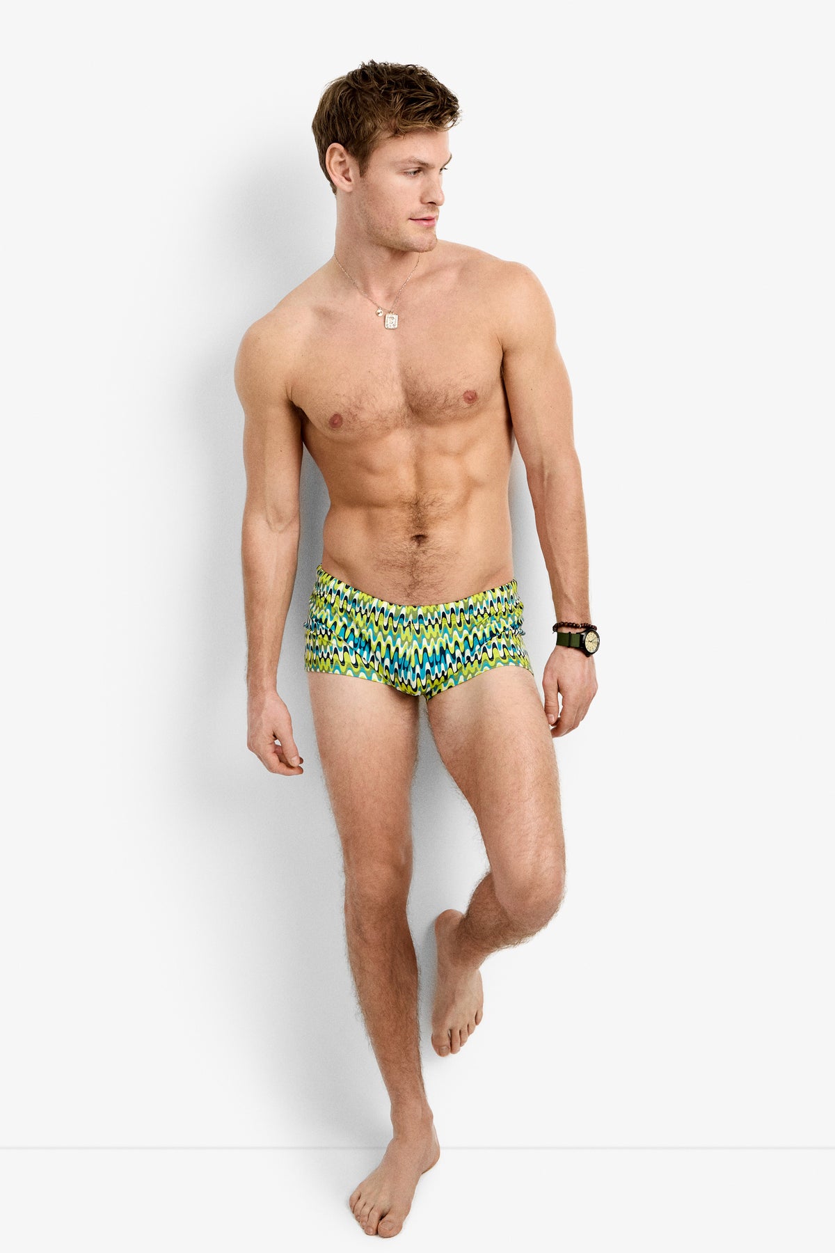 A shirtless man stands against a white background wearing the Sorrento Swim Trunk - Psychedelic Voyage, along with a necklace and wristwatch, with one leg slightly bent and one hand resting on the wall.