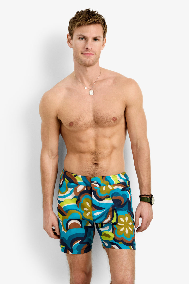 A shirtless man with short brown hair, a necklace, and a watch on his left wrist stands against a white background wearing Honolulu Board Short - Fonda Floral, featuring colorful blue, green, brown, and yellow prints—ideal summer swimwear.