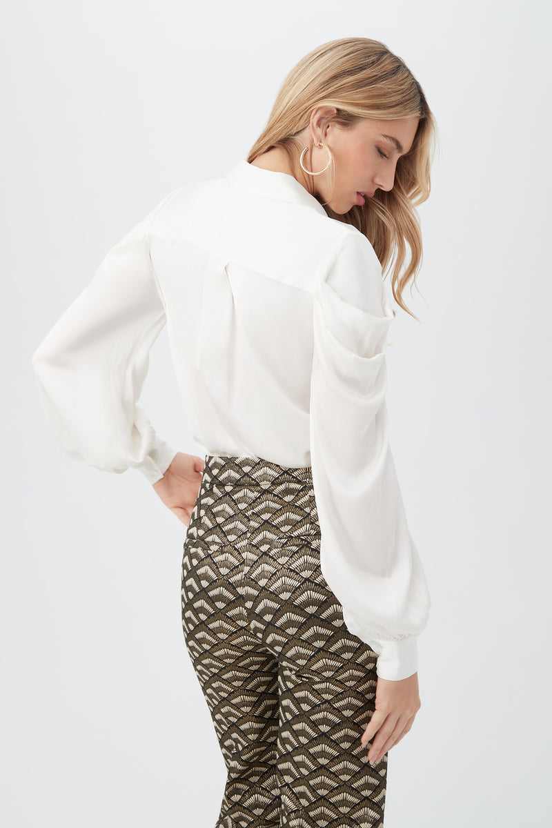A woman with long blonde hair poses with her back slightly turned, wearing the Bashira Top paired with high-waisted, patterned pants in black, gold, and whiteâ€â€showcasing an elegant and trendy look in women’s fashion.