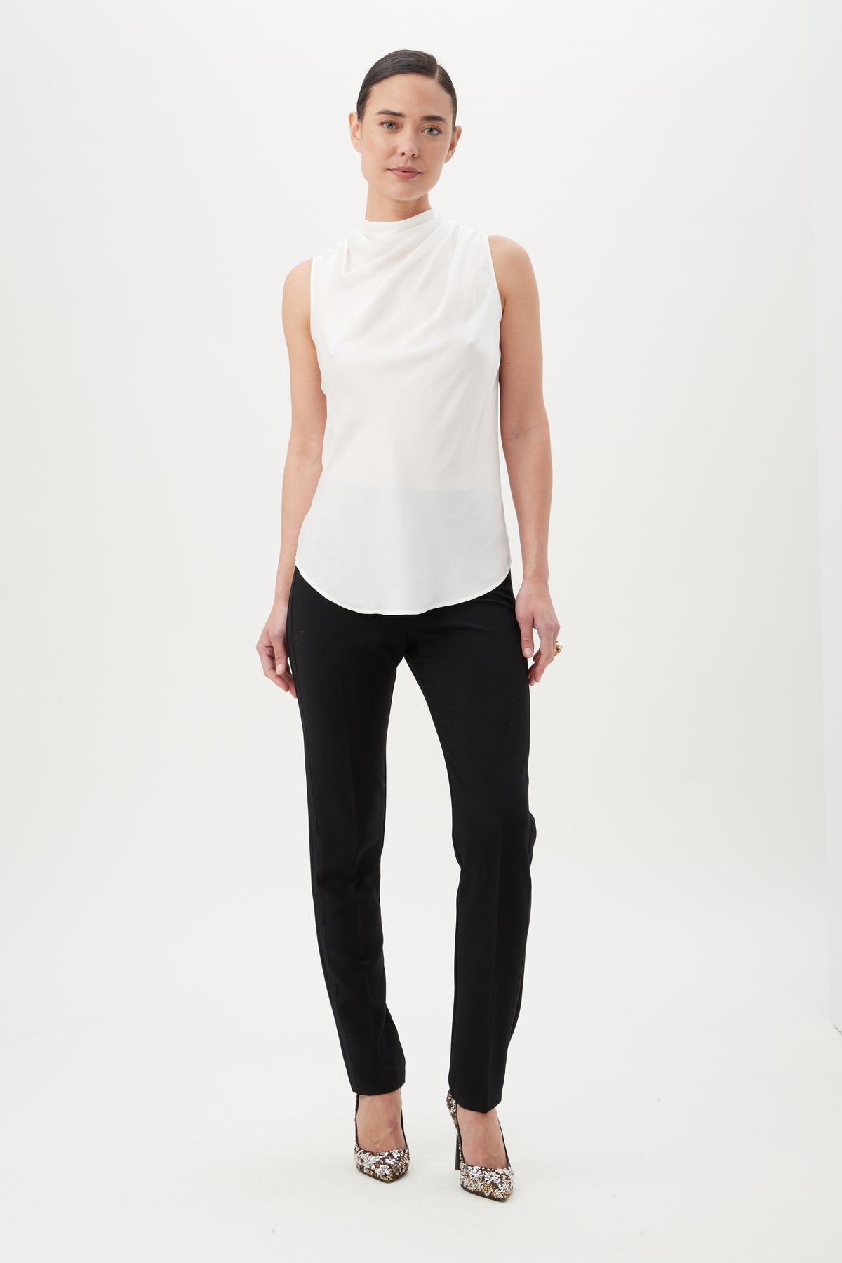 A woman stands against a white background wearing the sleeveless Lenaya Top with a funnel neckline, black pants, and patterned high heels. Her hair is pulled back and her arms rest relaxed at her sides.