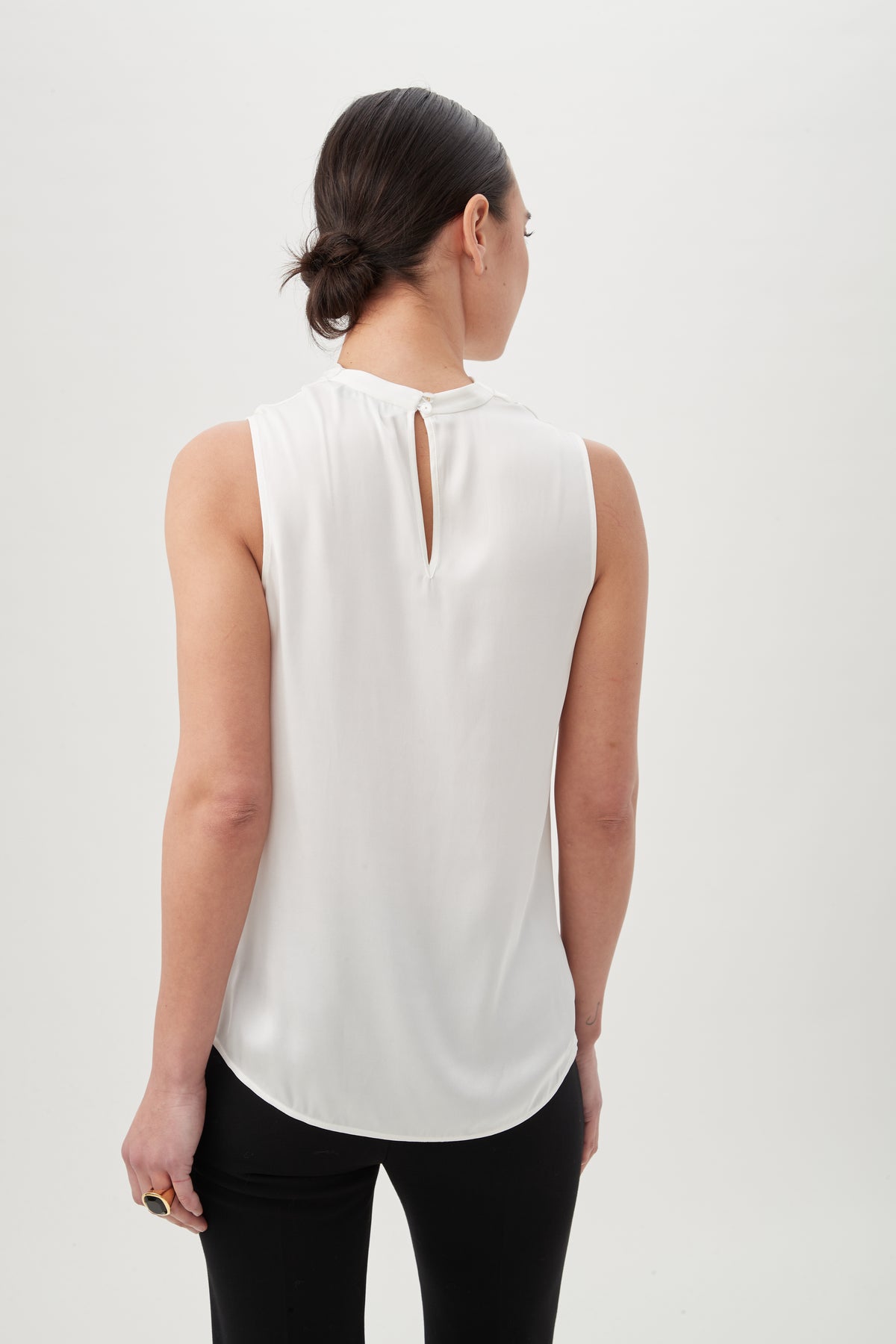 A woman with dark hair in a low bun is seen from behind wearing the Lenaya Top, a sleeveless stretch satin style with a small back keyhole, paired with black pants against a plain white background.