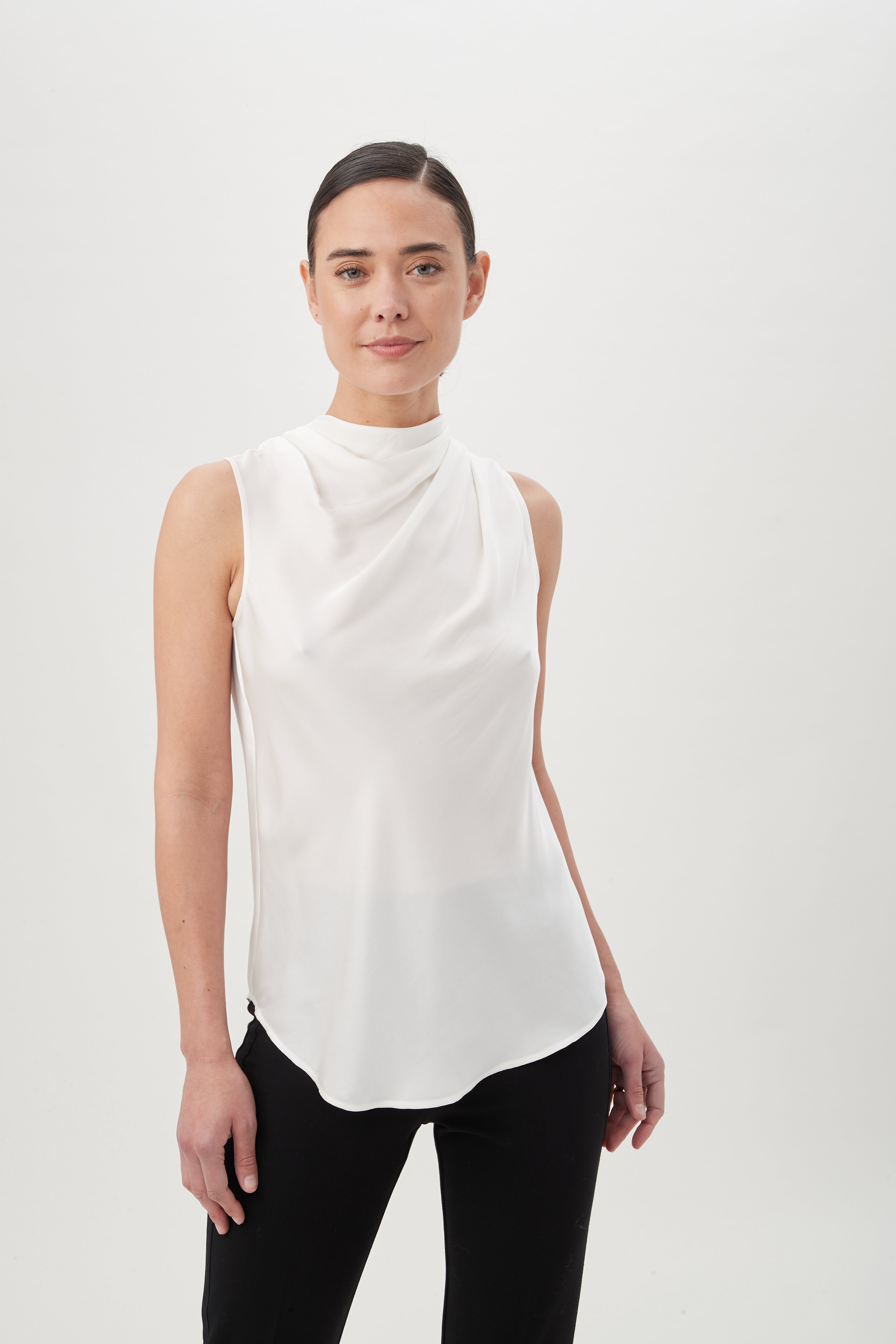 A person stands against a plain white background, wearing the Lenaya Top—a sleeveless, funnel-neck top—paired with black pants. Their dark hair is pulled back as they look slightly to the side with a neutral expression.