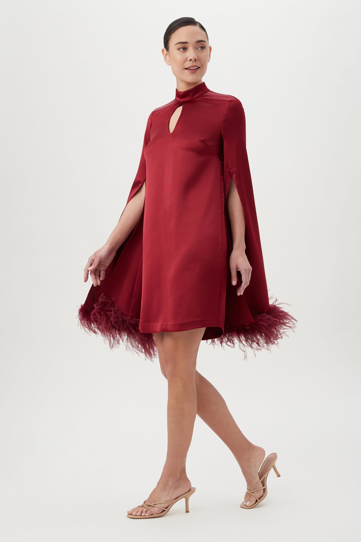 A woman in the Hiromi 2 Dressâ€â€red satin, sheath silhouette, keyhole neckline, and feather-trimmed hemâ€â€walks in beige heeled sandals against a plain white background.