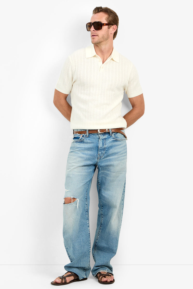 Man wearing sunglasses, an Asher Polo (men’s slim fit Merino Wool), distressed blue jeans with knee rip, brown belt, and sandals stands with hands in pockets against a plain background.