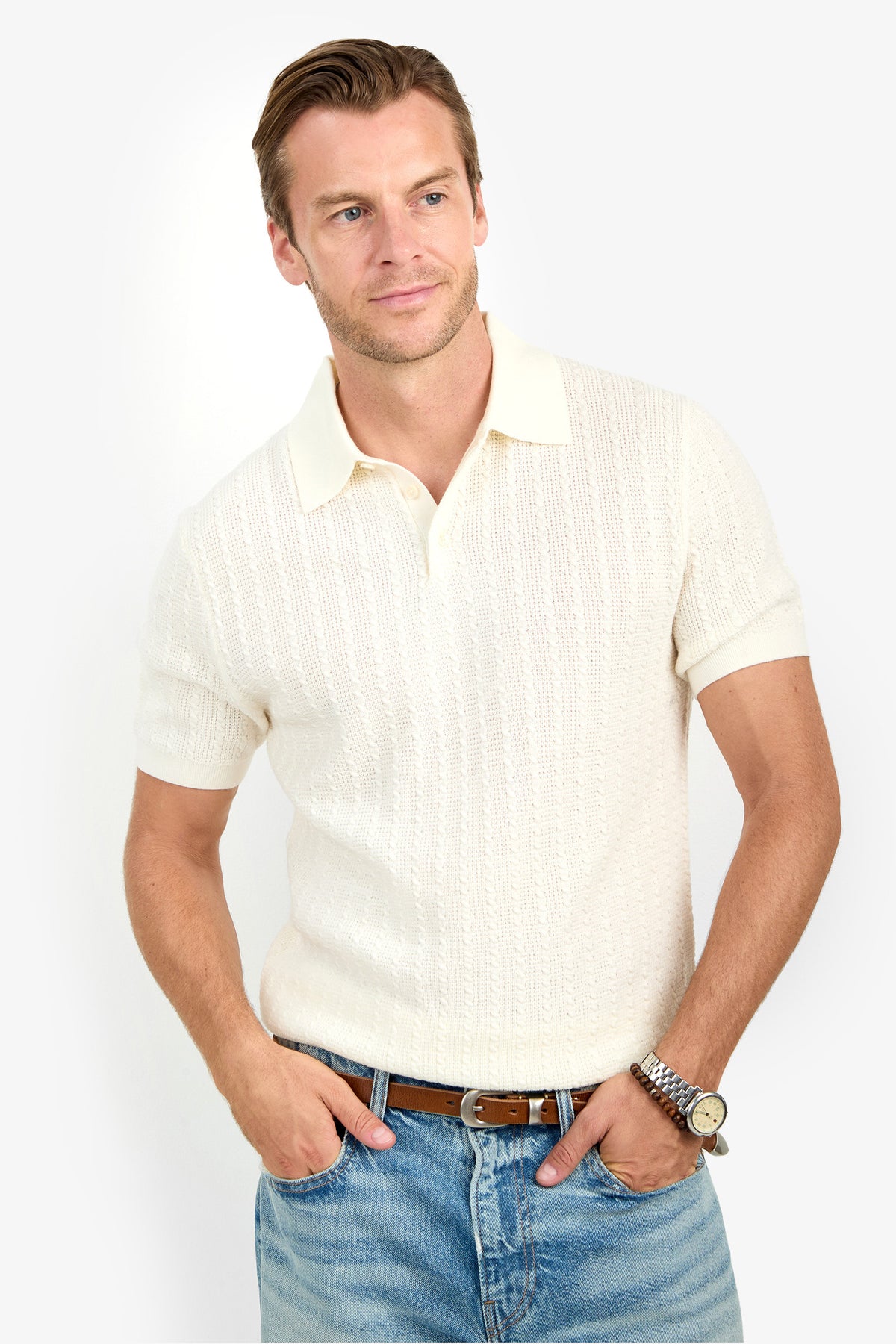 A man wearing the Asher Polo in cream and light blue jeans stands with hands in pockets, looking to the side and smiling.