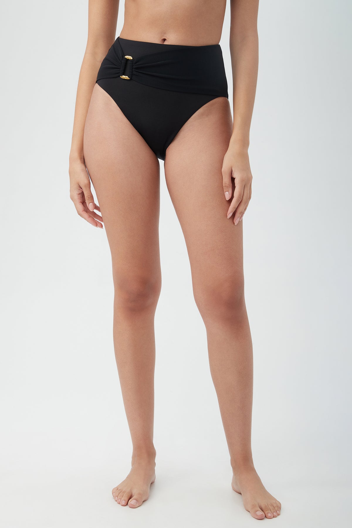 A person models the Women's Monaco Belted High Waist Swim Bottom in black, featuring a gold buckle detail, standing barefoot against a plain light background with arms relaxed at their sides.