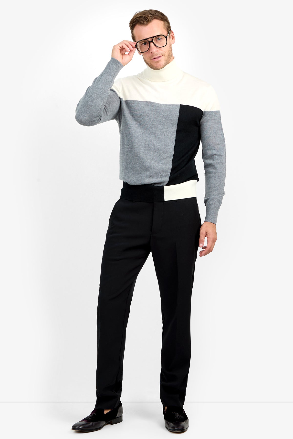 A man wearing glasses, the Callisto Sweater in white, gray, and black, and black pants stands against a plain white background, holding his glasses with one hand.