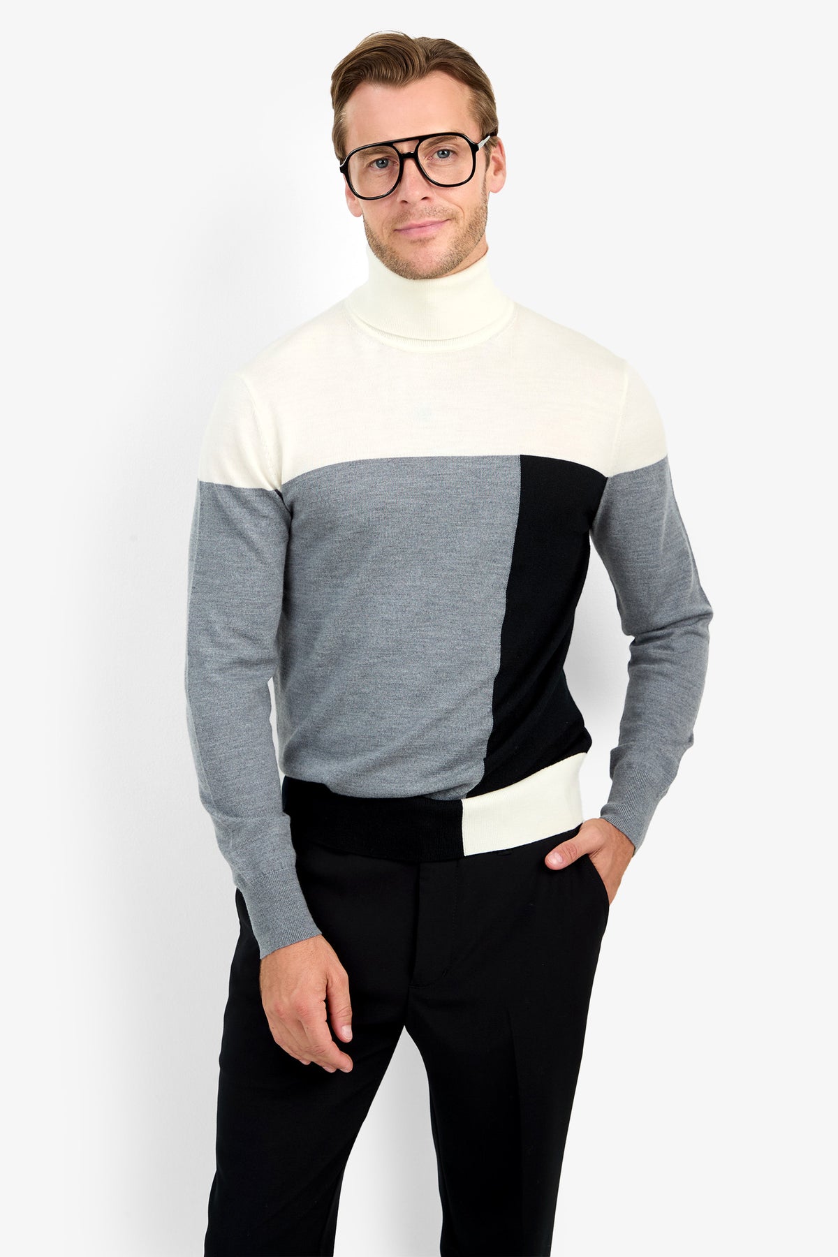 Man wearing large glasses, the Callisto Sweater in white, gray, and black color-blocking, and black pants stands with one hand in his pocket against a plain white background.