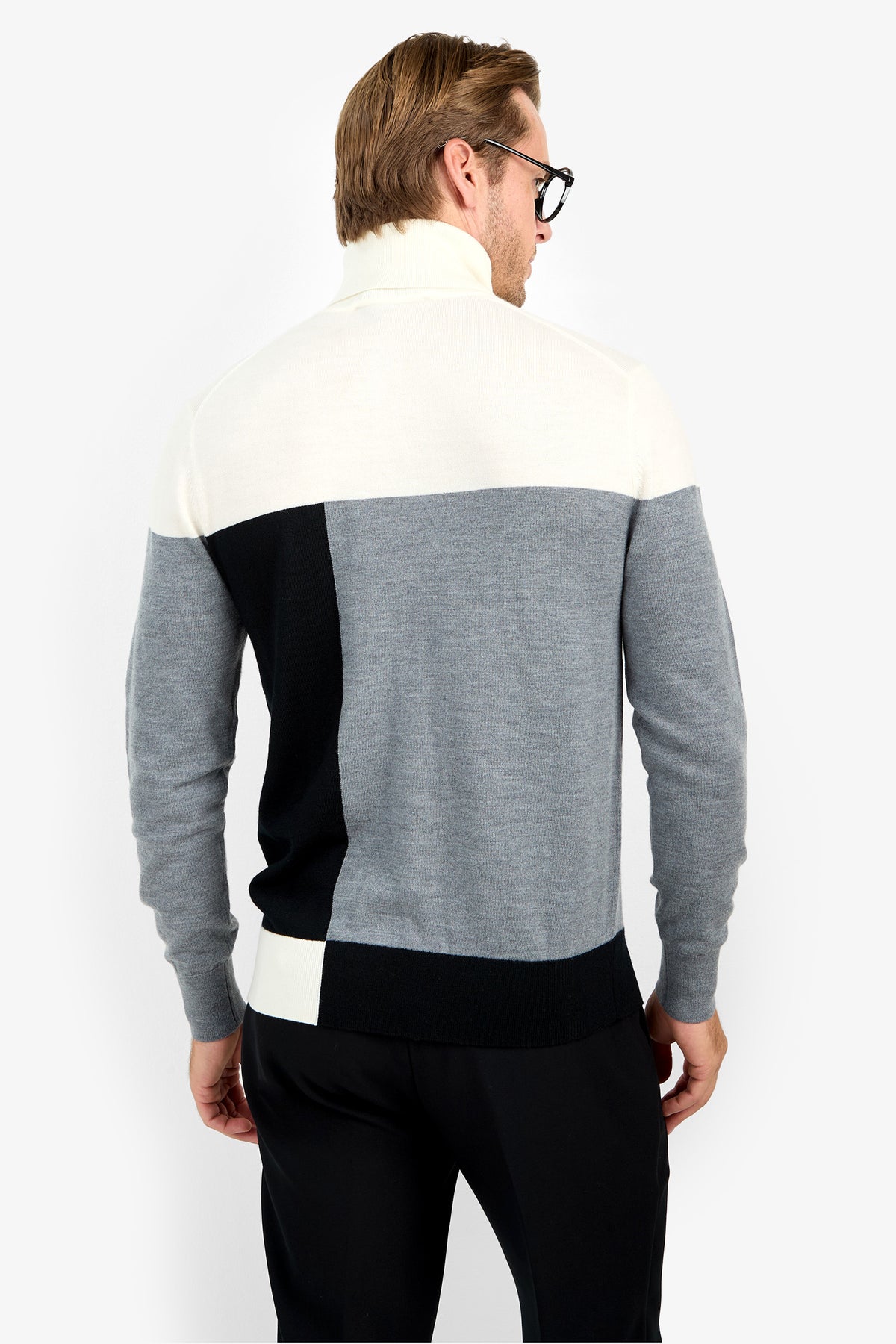 A man in glasses and the Callisto Sweater—a color-blocked merino wool design in white, gray, and black—stands with his back to the camera against a plain white background.
