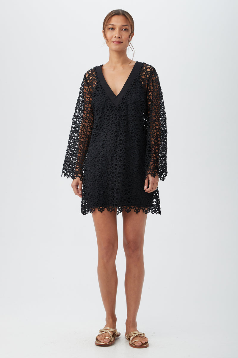 A woman with her hair tied back stands against a plain background, wearing the Women's Chateau Lace Long Sleeve V-Neck Swim Cover-Up Dress in black and tan sandals, displaying a neutral expression.
