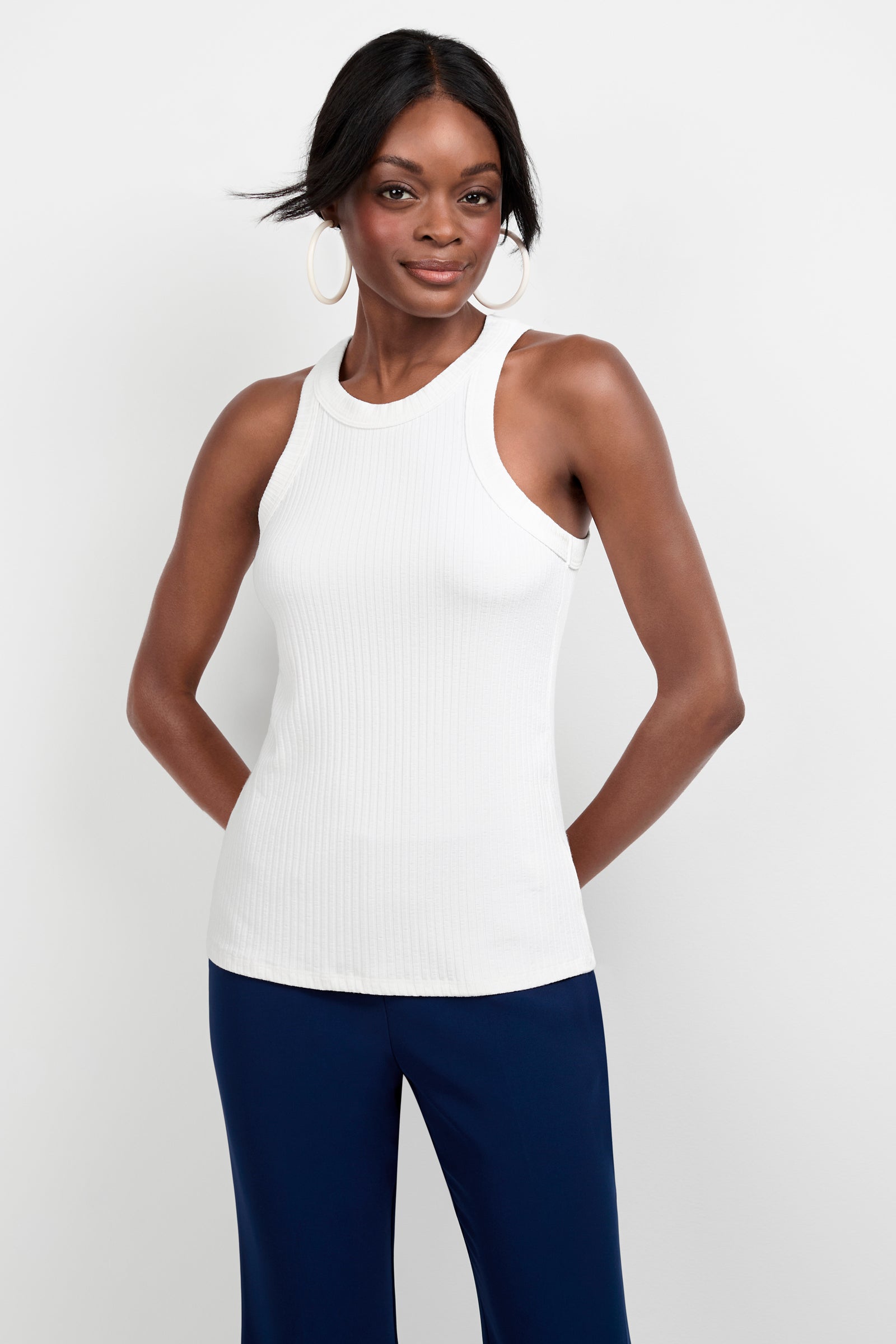 A woman with dark skin and straight black hair wears large hoop earrings, navy blue pants, and a sleeveless white Continental Tank Top. She stands against a plain white background, smiling with her hands behind her back.