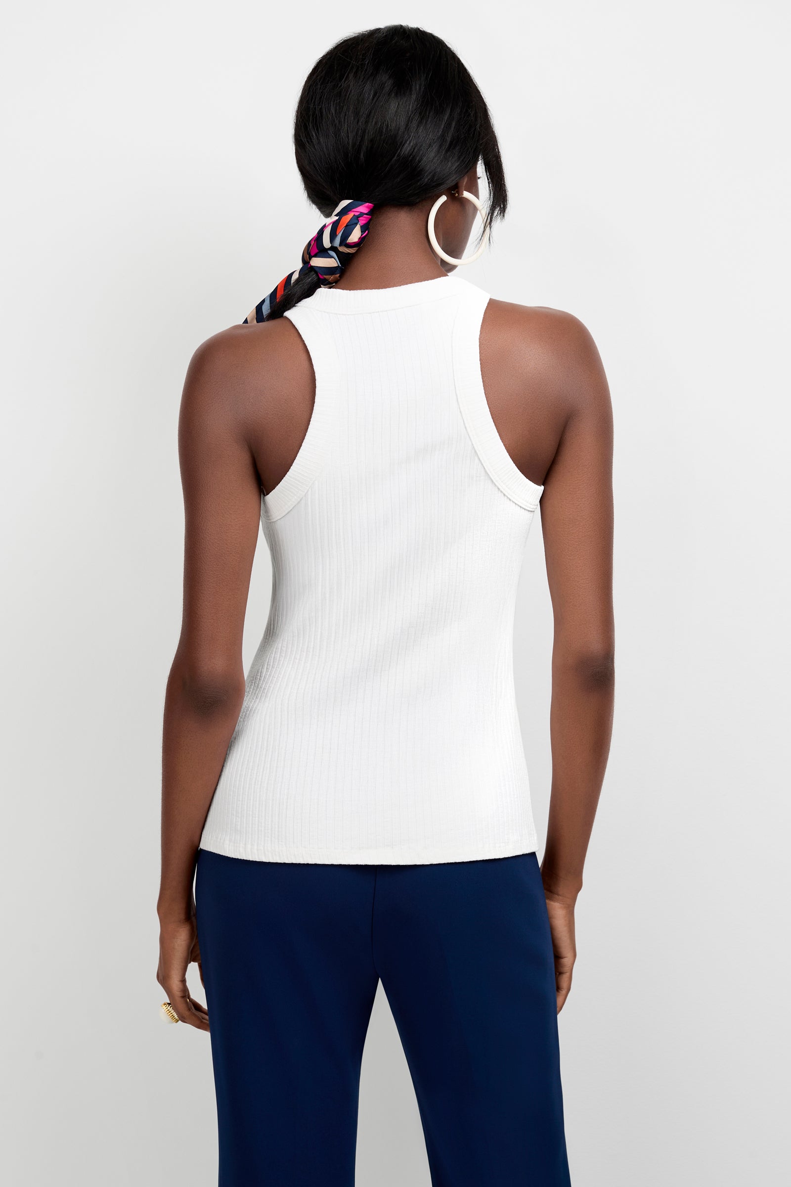 A woman is shown from the back wearing the Continental Tank Top in white, navy pants, large hoop earrings, and a colorful scarf tied around her hair against a plain white background.
