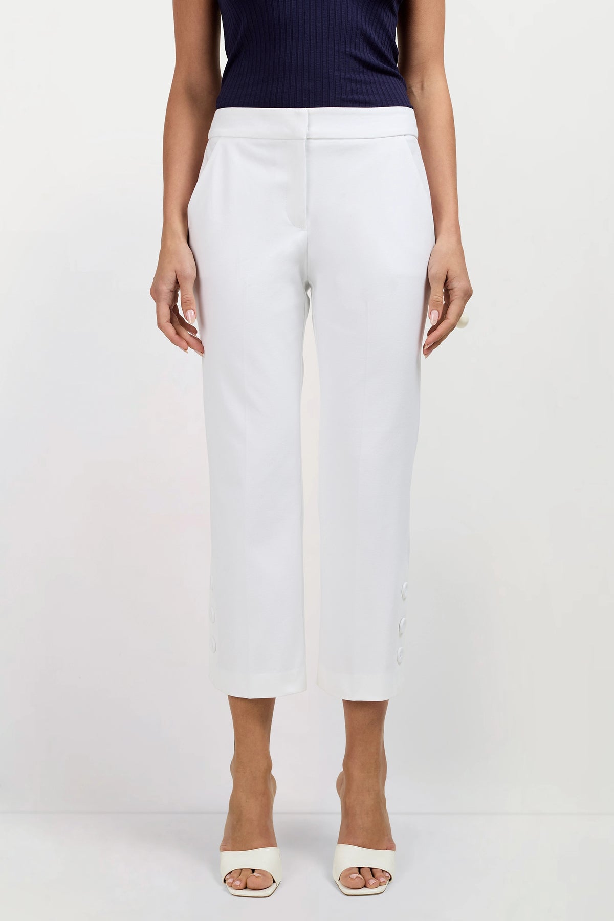A person in Ore Button-Hem Cropped Pants, styled with a dark sleeveless top and white open-toed heels, stands against a plain light background.