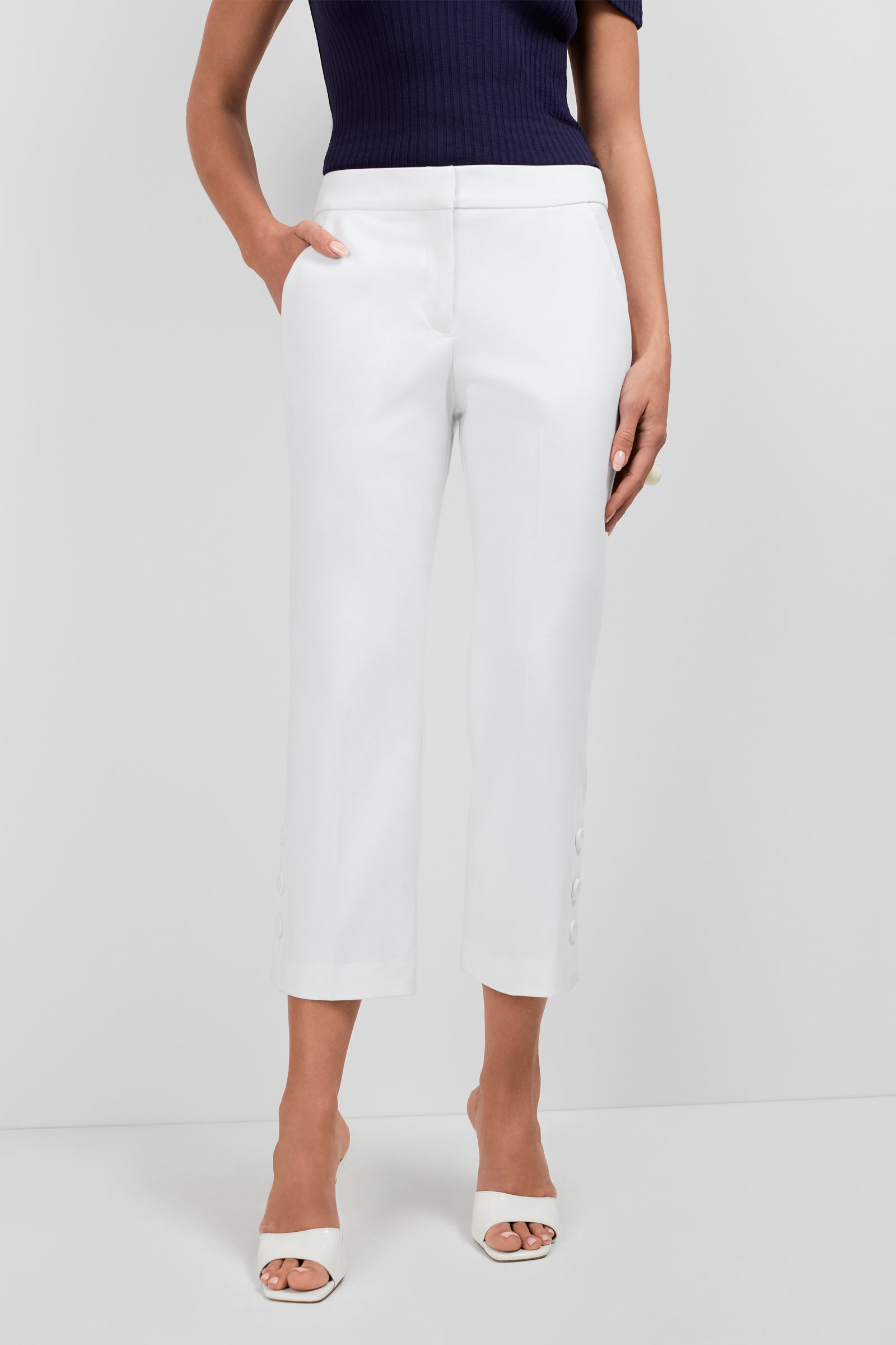 Someone stands against a plain light background wearing Ore Button-Hem Cropped Pants in white, featuring button details at the ankles, paired with a dark ribbed short-sleeve top and white open-toe heels.