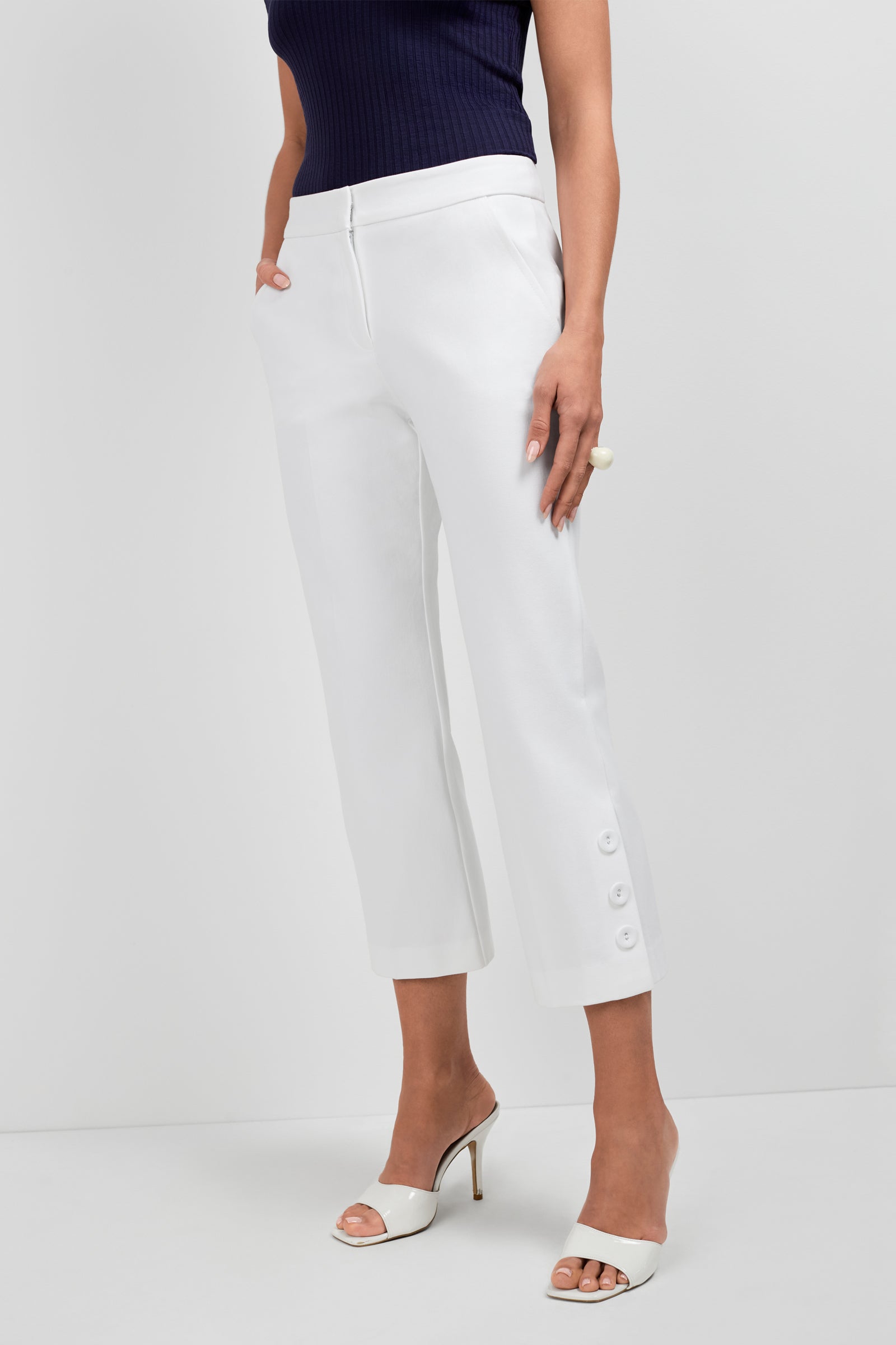 A woman in Ore Button-Hem Cropped Pants, a sleeveless navy top, white heeled sandals, and a large ring stands against a plain light background.