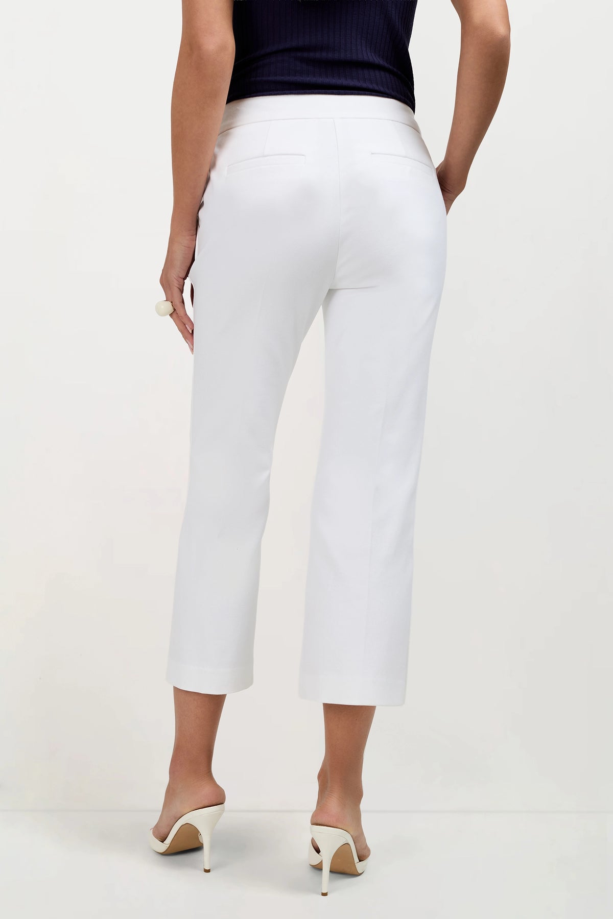 A woman stands facing away, wearing Ore Button-Hem Cropped Pants, a sleeveless black top, and white heeled sandals against a plain white background.