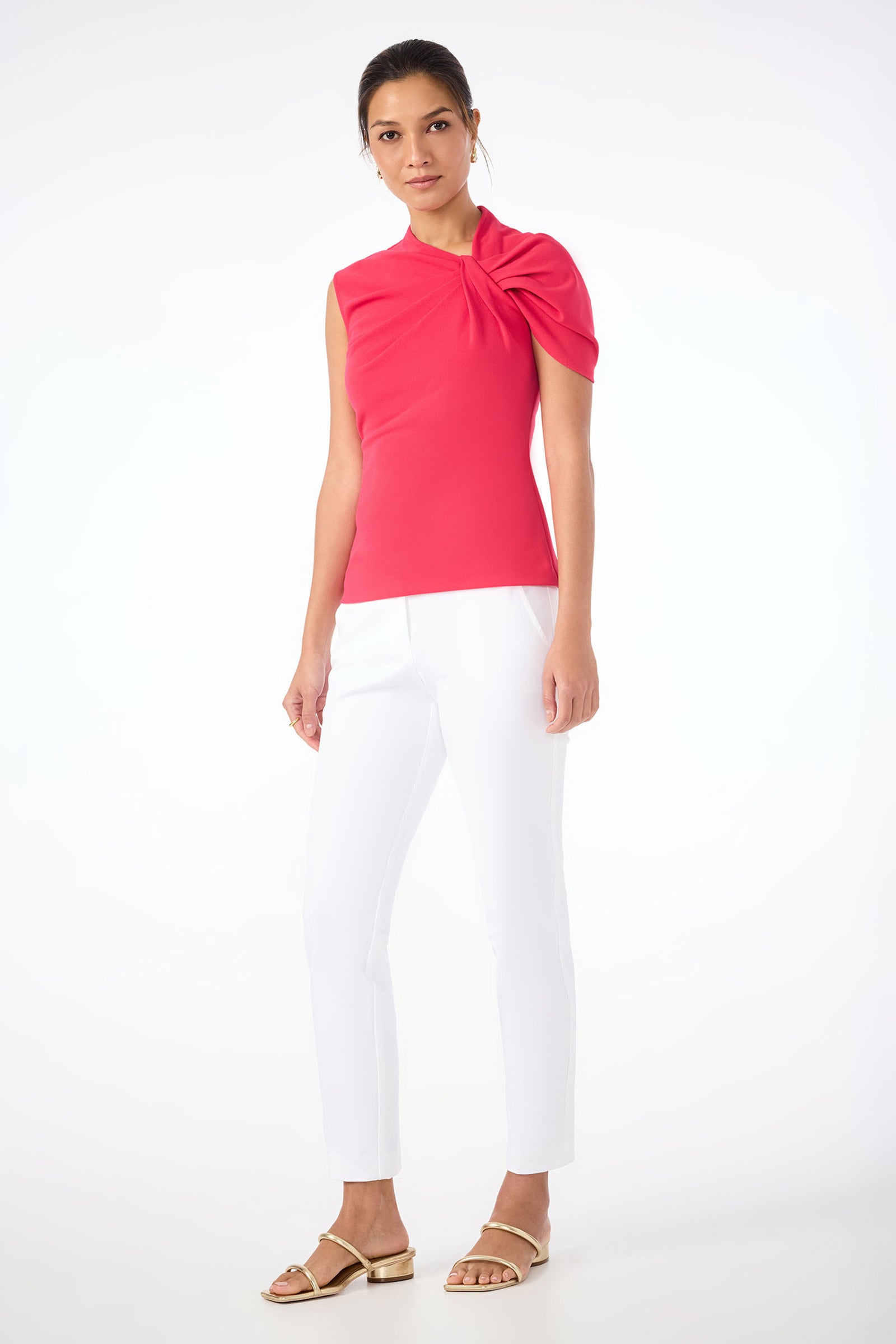 A woman stands against a white background wearing white Aubree 2 Cigarette Pants, a bright pink asymmetrical top with gathered detail on one shoulder, and gold flat sandals.
