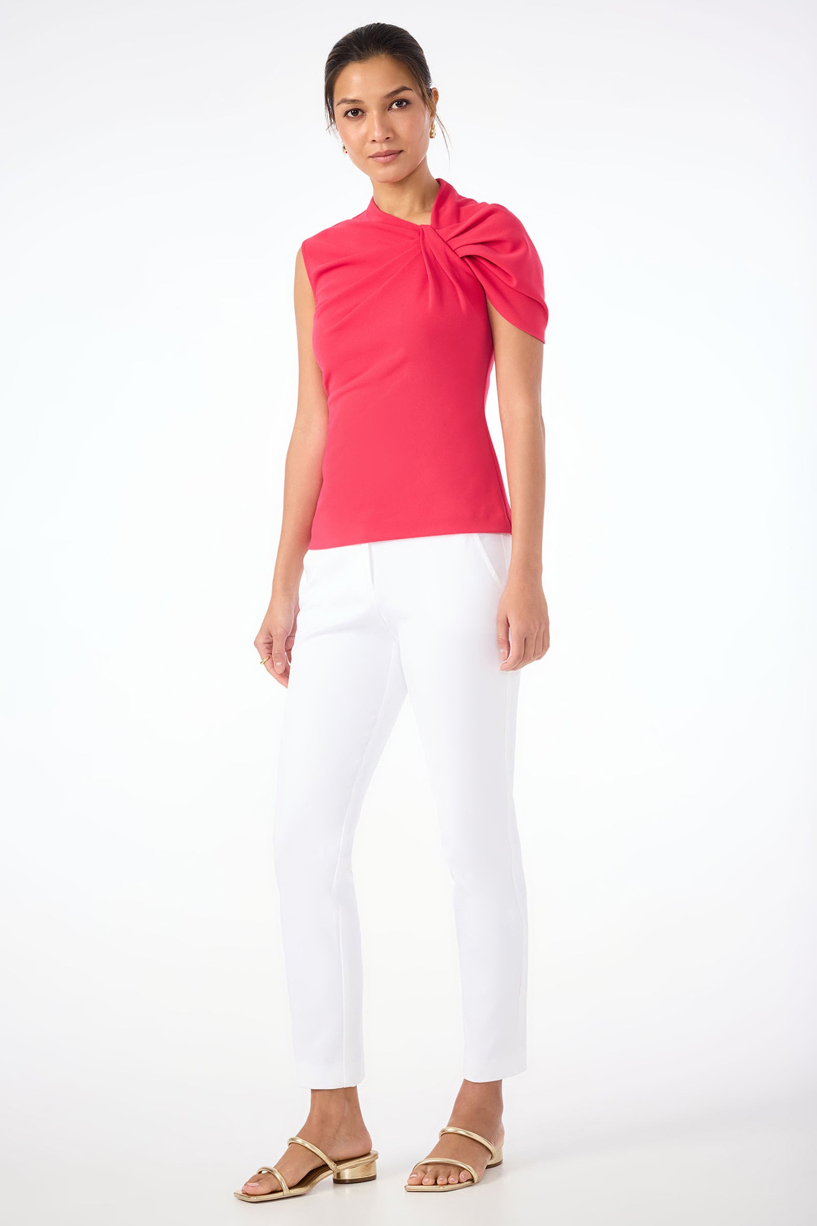 A woman stands against a white background wearing the Keshi Short-Sleeve Twist Top in bright pink, styled with white slim-fit pants and beige sandals.