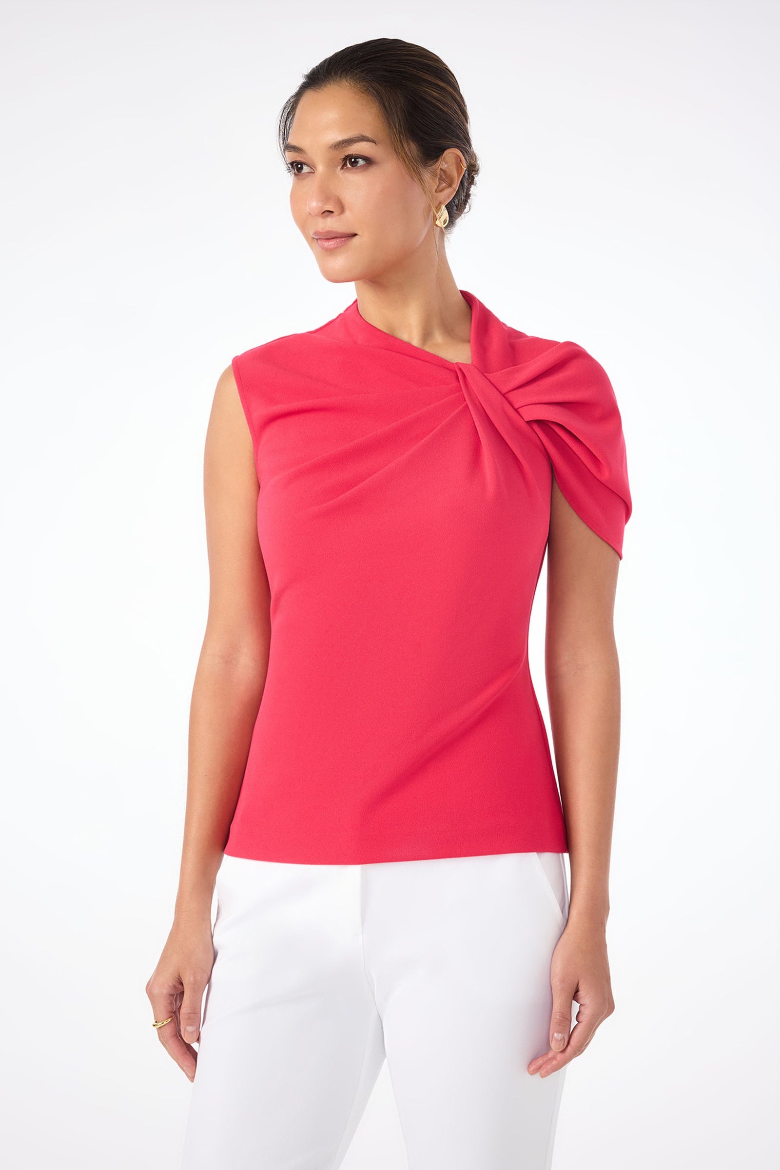 A woman wears the Keshi Short-Sleeve Twist Top in bright pink, featuring a gathered detail on one shoulder, paired with white pants and standing against a plain white background.