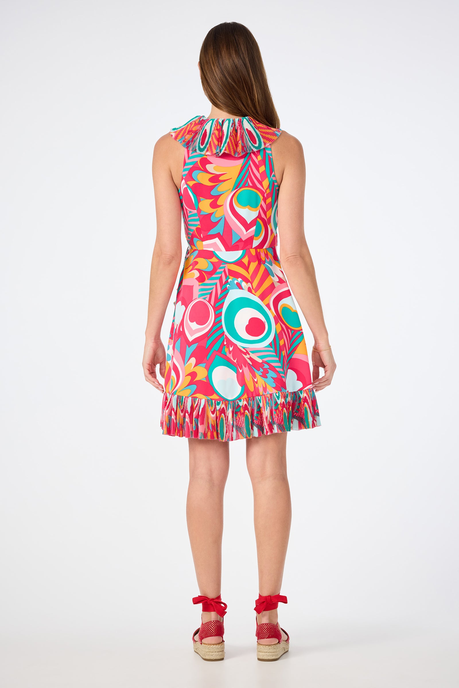 A woman faces away, wearing the Marseille Sleeveless Wrap Dress - Dramont, featuring a bold red, pink, green, and white abstract pattern and a ruffled hem. She pairs it with red wedge sandals.