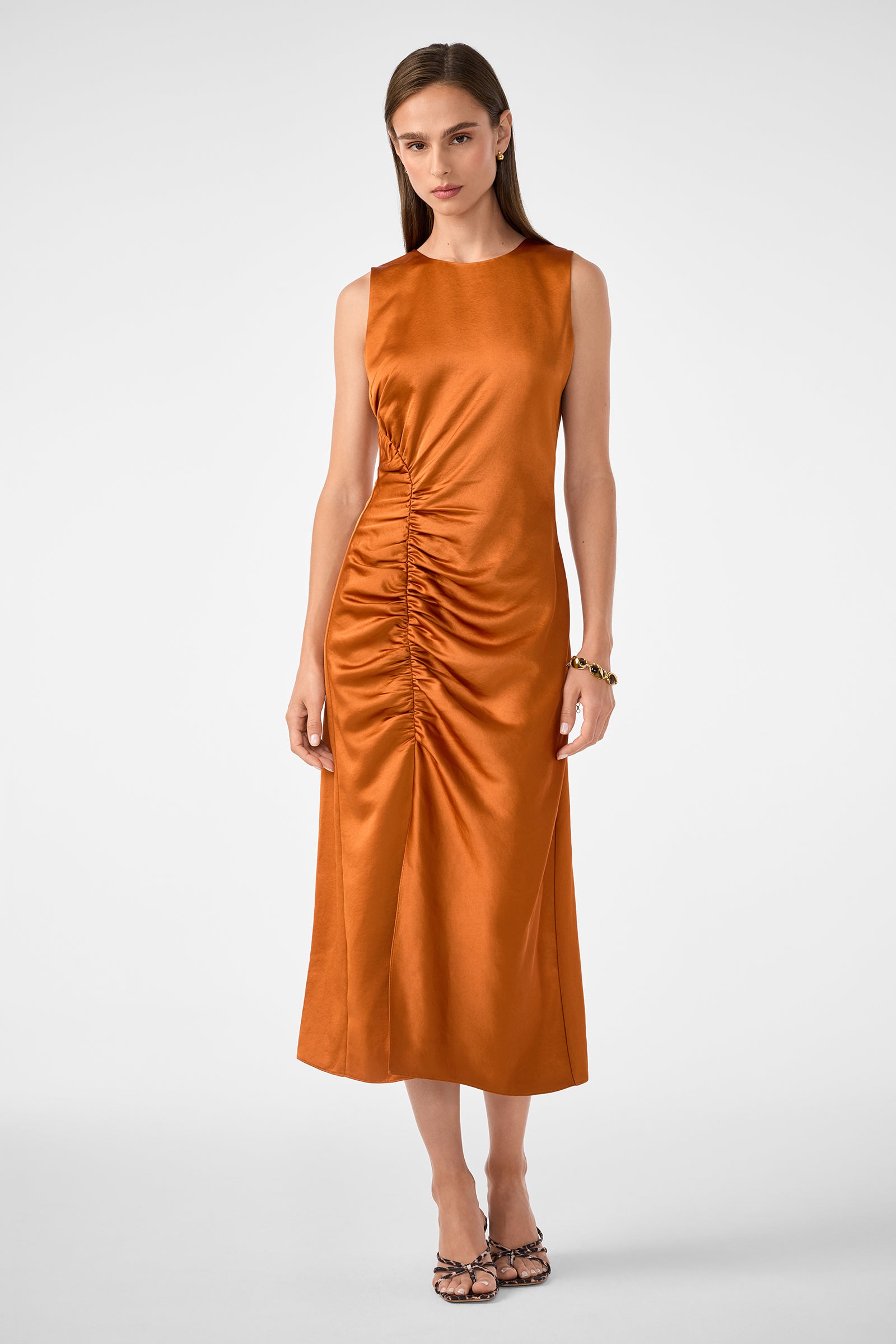 A woman stands against a plain background wearing the Chestnut Dress—a sleeveless, shiny rust-orange midi with ruched side detailing—paired with strappy heeled sandals and a bracelet.