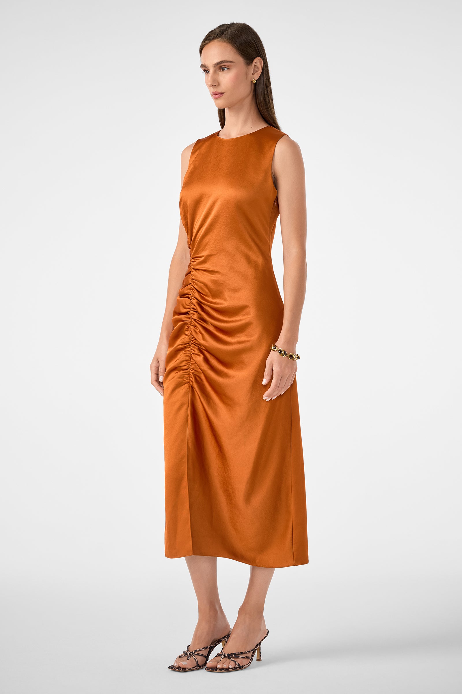 A woman wearing the Chestnut Dress, a sleeveless rust-orange midi with front ruching, stands in heels against a neutral background.