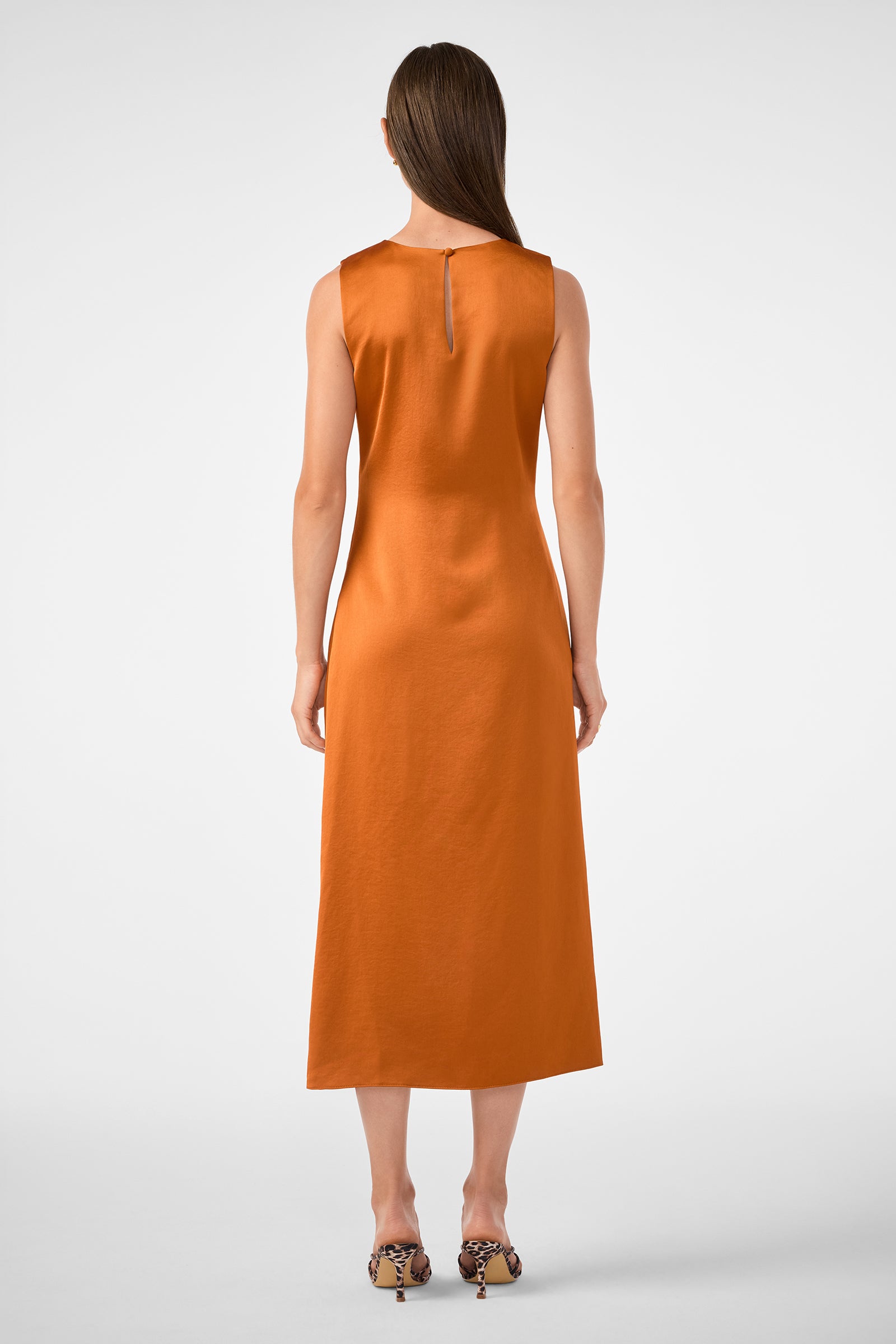 A woman faces away, wearing the sleeveless, ankle-length Chestnut Dress in burnt orange with patterned heeled sandals. Her straight brown hair falls down her back against a plain white background.