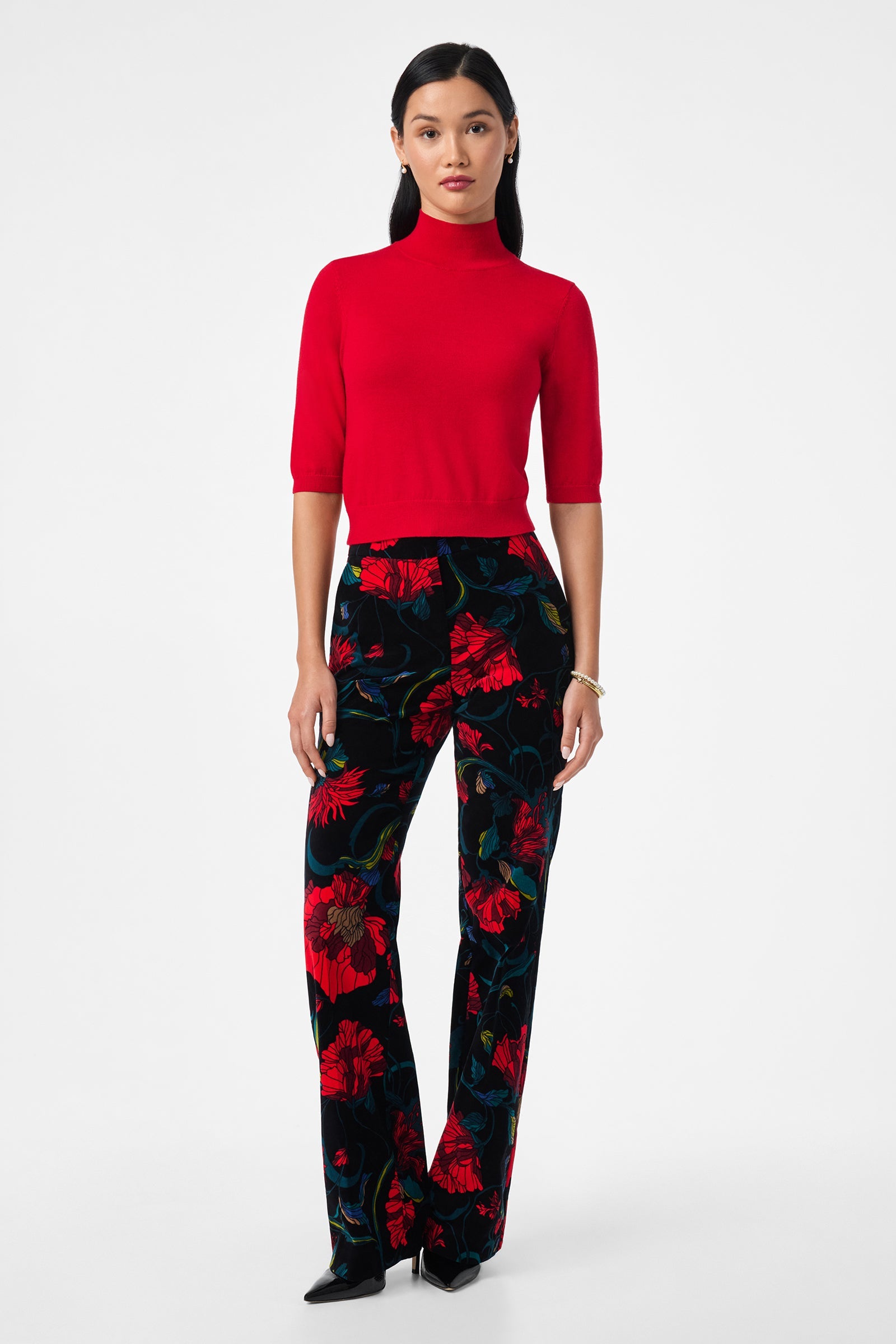 A woman with straight dark hair and minimal jewelry stands against a plain background wearing the fitted red Mali Mock Neck top, black pants with bold red floral print, and black shoes.
