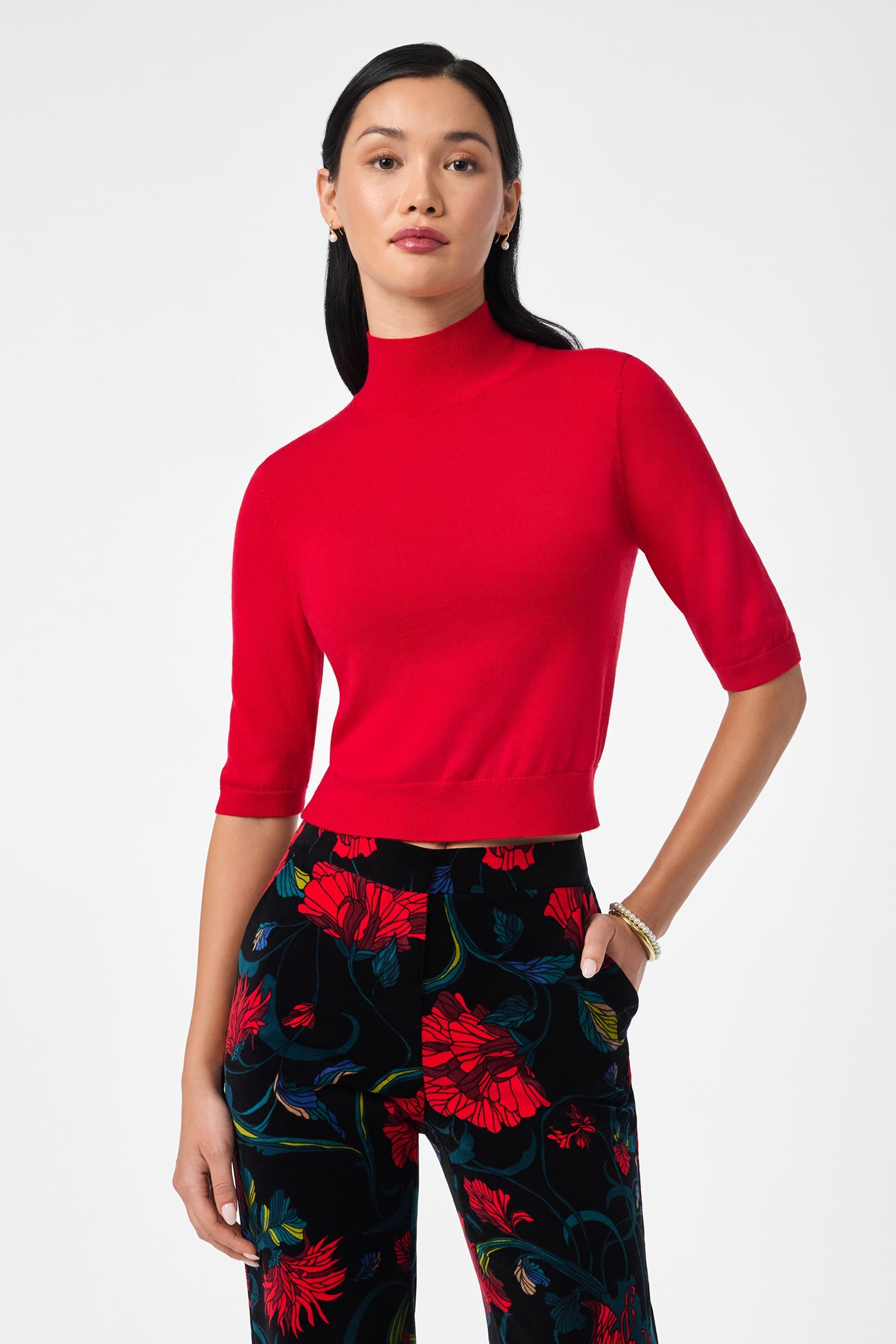 A woman wears the Mali Mock Neck in Moulin Rouge and black pants with a red and blue floral pattern, standing against a plain white background.