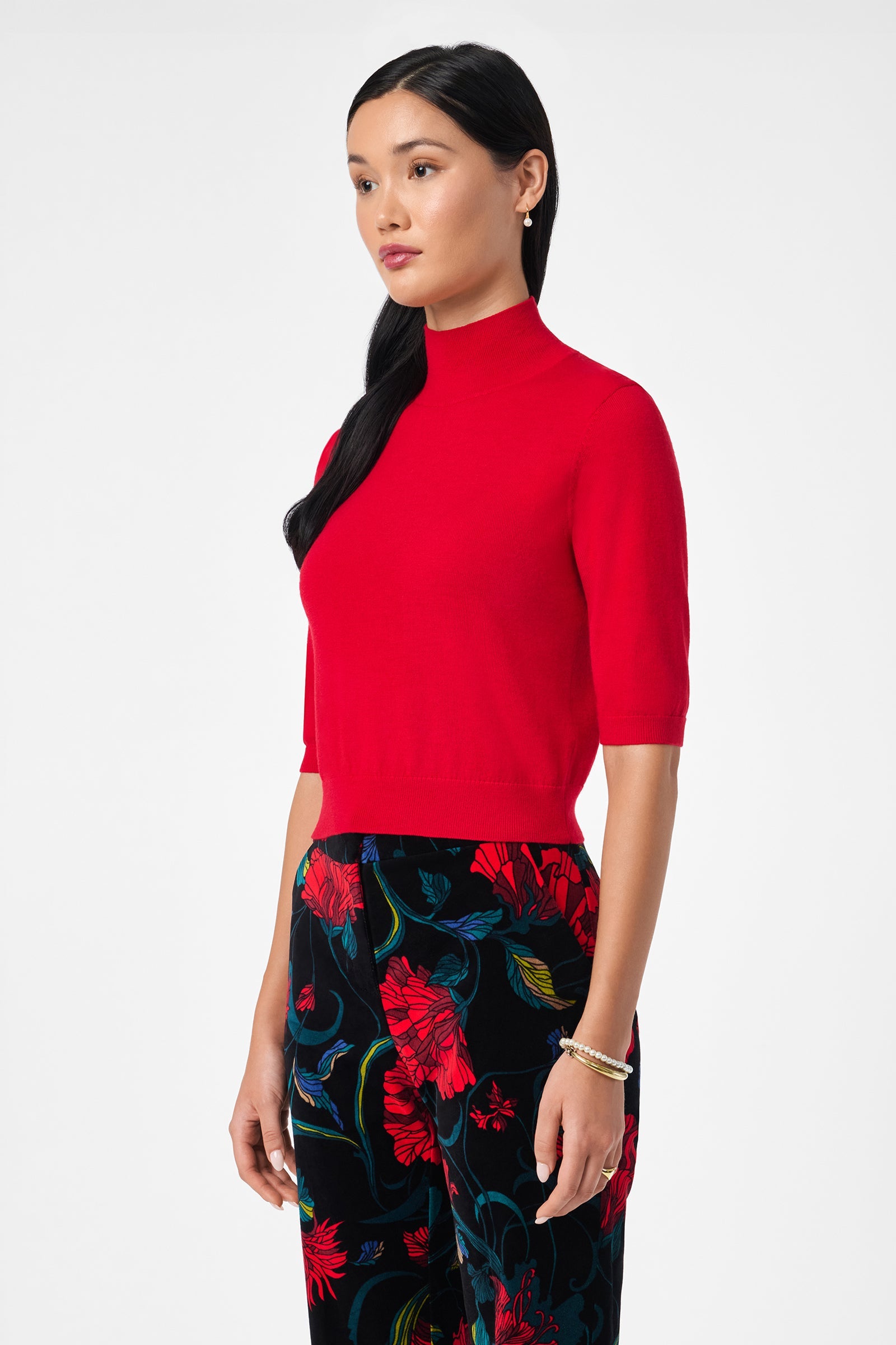 A woman with long black hair wears the Mali Mock Neck in bright red and black floral pants with bold red and blue patterns. She stands against a plain white background, looking slightly to the side.