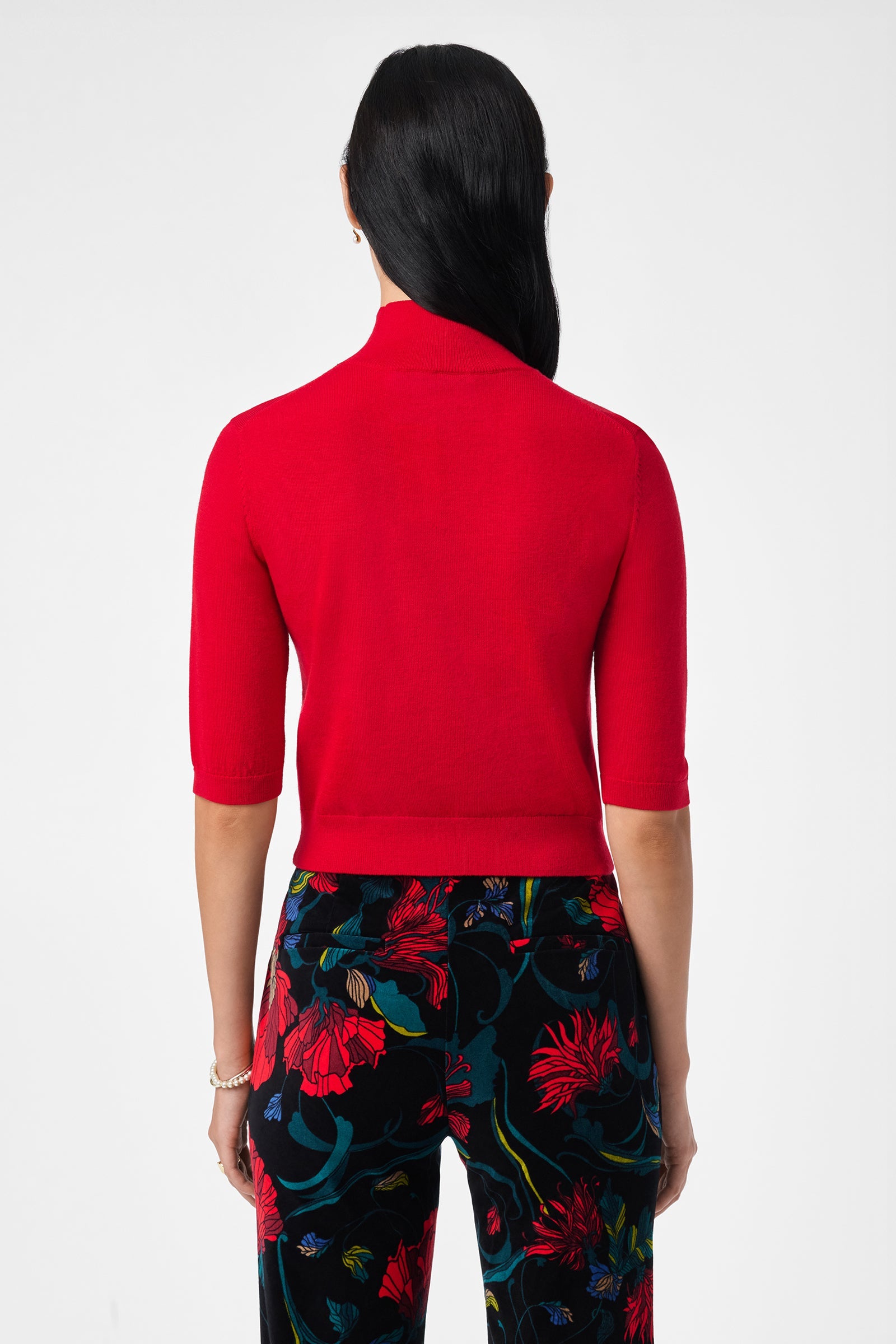 A woman is seen from the back wearing the bright red Mali Mock Neck top and high-waisted black pants with a vibrant floral pattern. Her straight dark hair falls down her back.