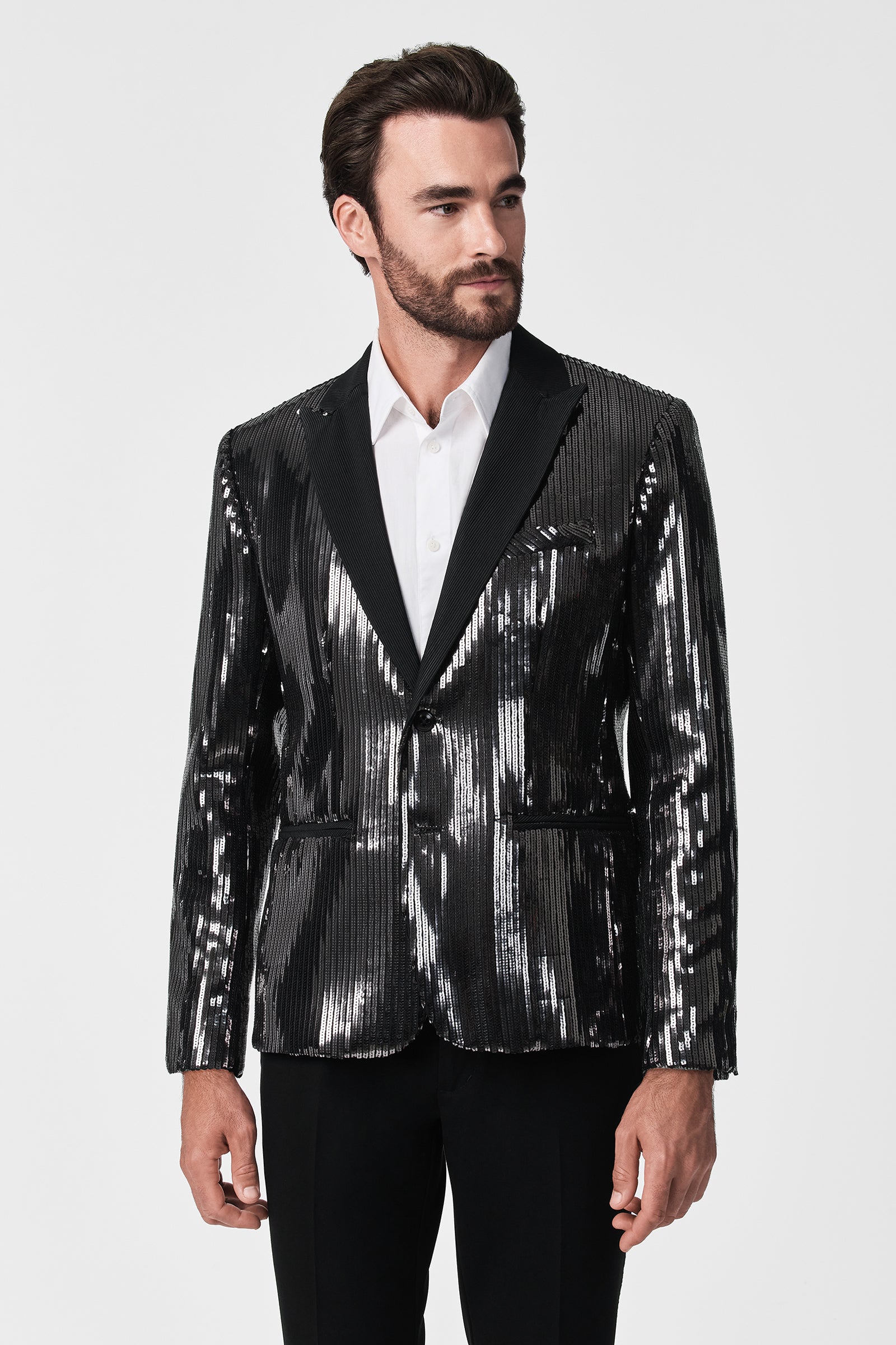 A man with dark hair and a beard models the shiny black Coleman Blazer over a white dress shirt and black pants, standing against a plain white background.