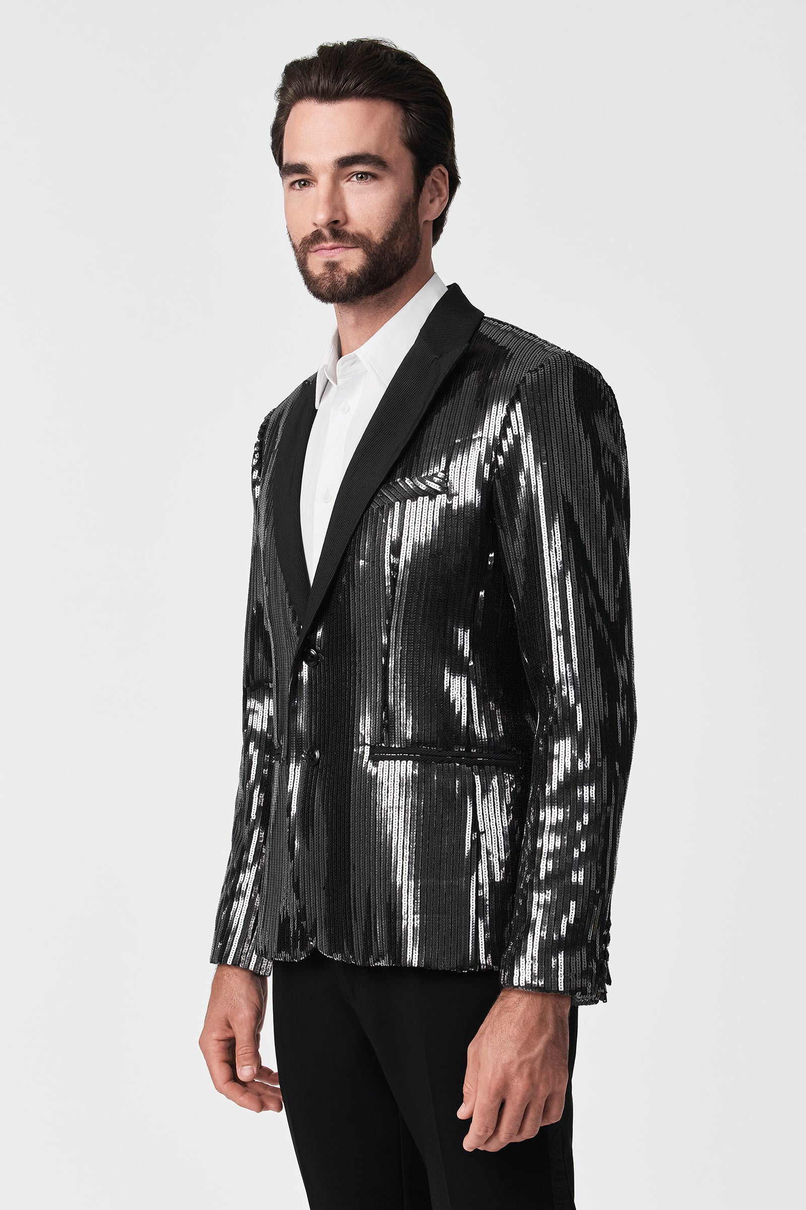 A man with dark hair and a beard wears the Coleman Blazer—a shiny black blazer with metallic stripes—over a white dress shirt and black pants, posing against a plain white background.