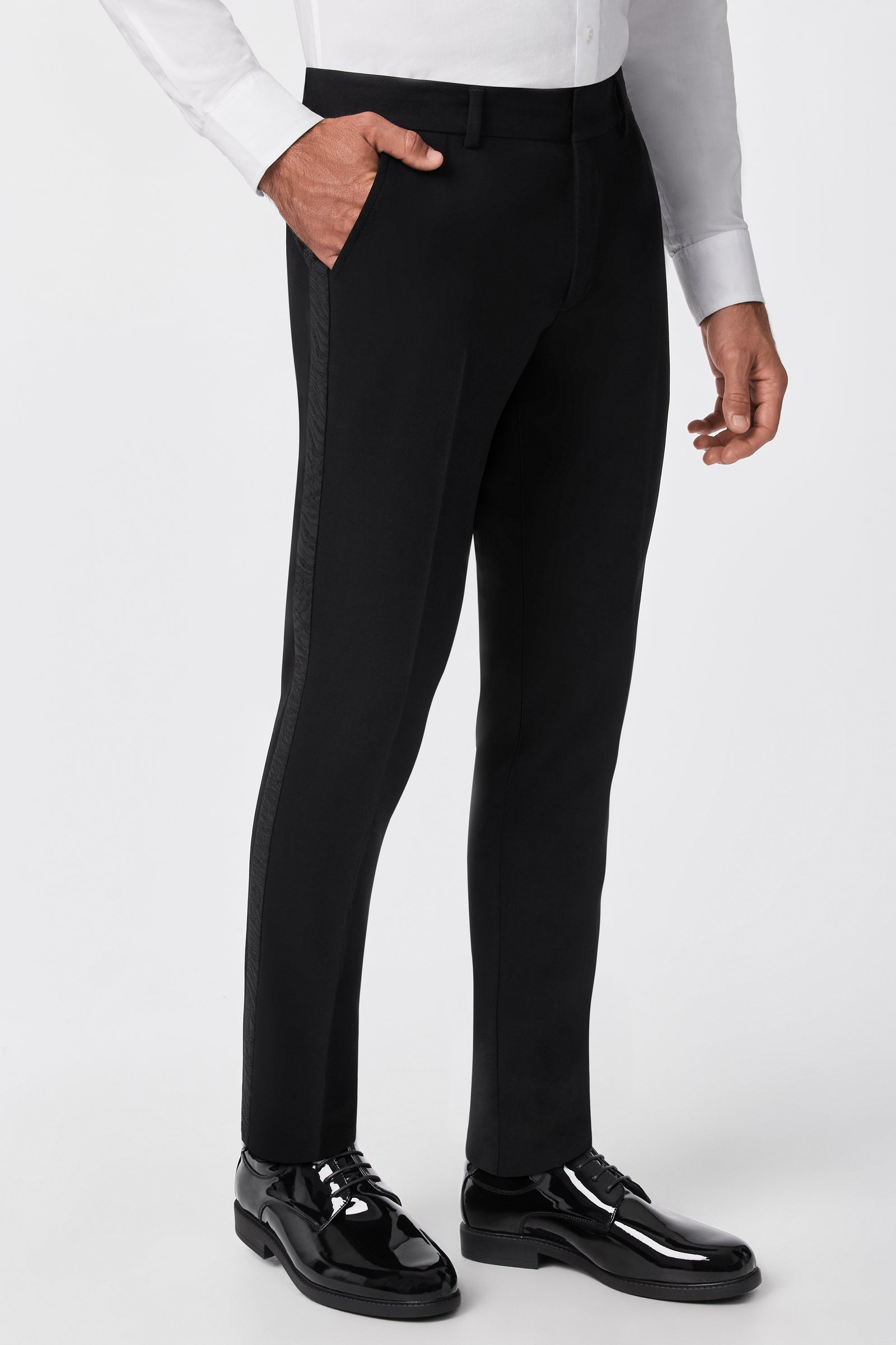 A man stands against a plain background with one hand in his pocket, wearing the Adrian Tux Trouser featuring a satin side stripe, paired with a white dress shirt and black shiny dress shoes.