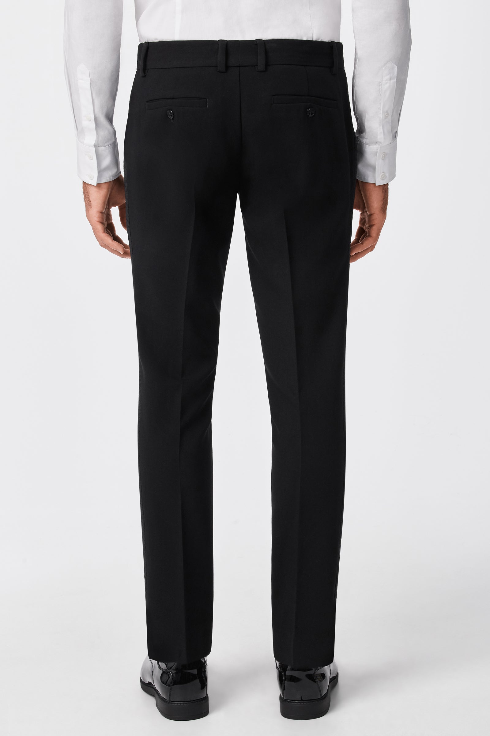 A person is shown from the back wearing the Adrian Tux Trouser, featuring belt loops and buttoned back pockets, paired with a white dress shirt against a plain white background.