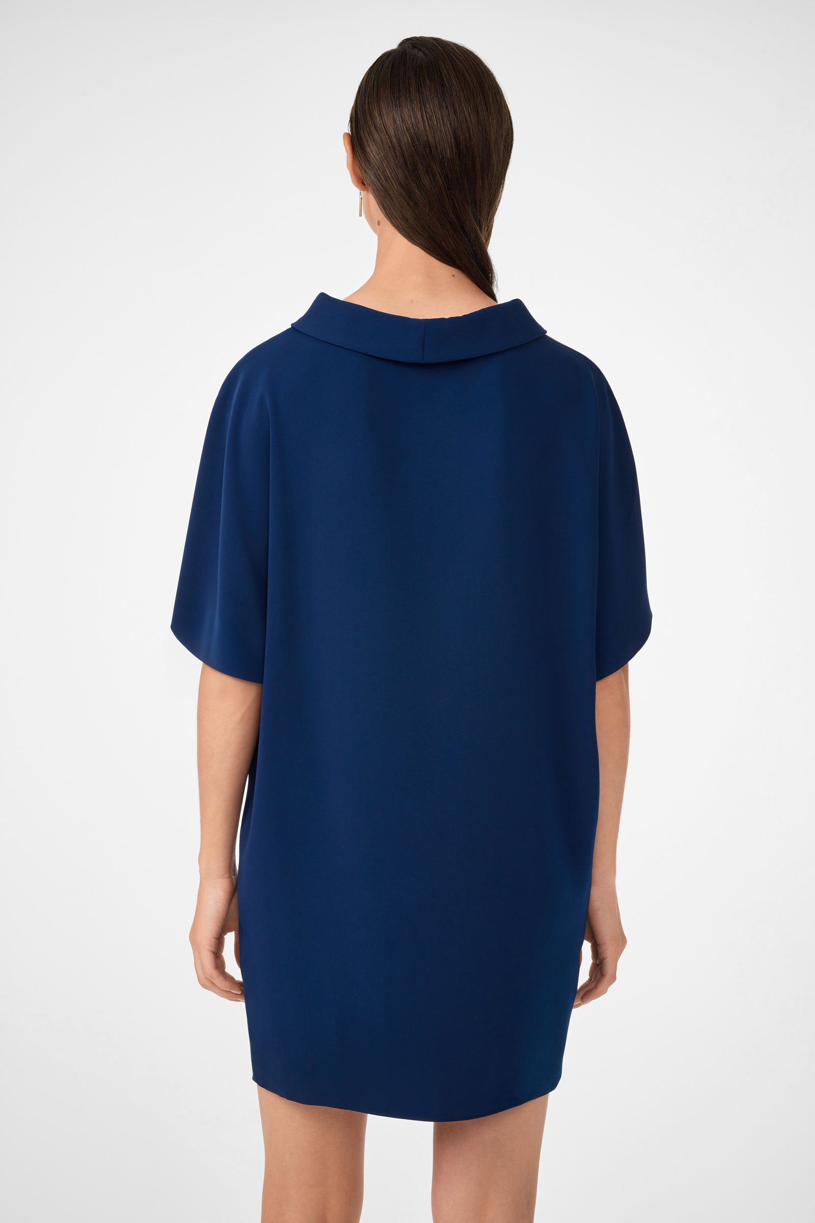 A woman with long brown hair in a low ponytail is shown from the back, modeling the Honeysuckle Short Dolmain Sleeve Shift Dress—a loose-fitting, short-sleeved, knee-length dark blue dress—against a plain white background.