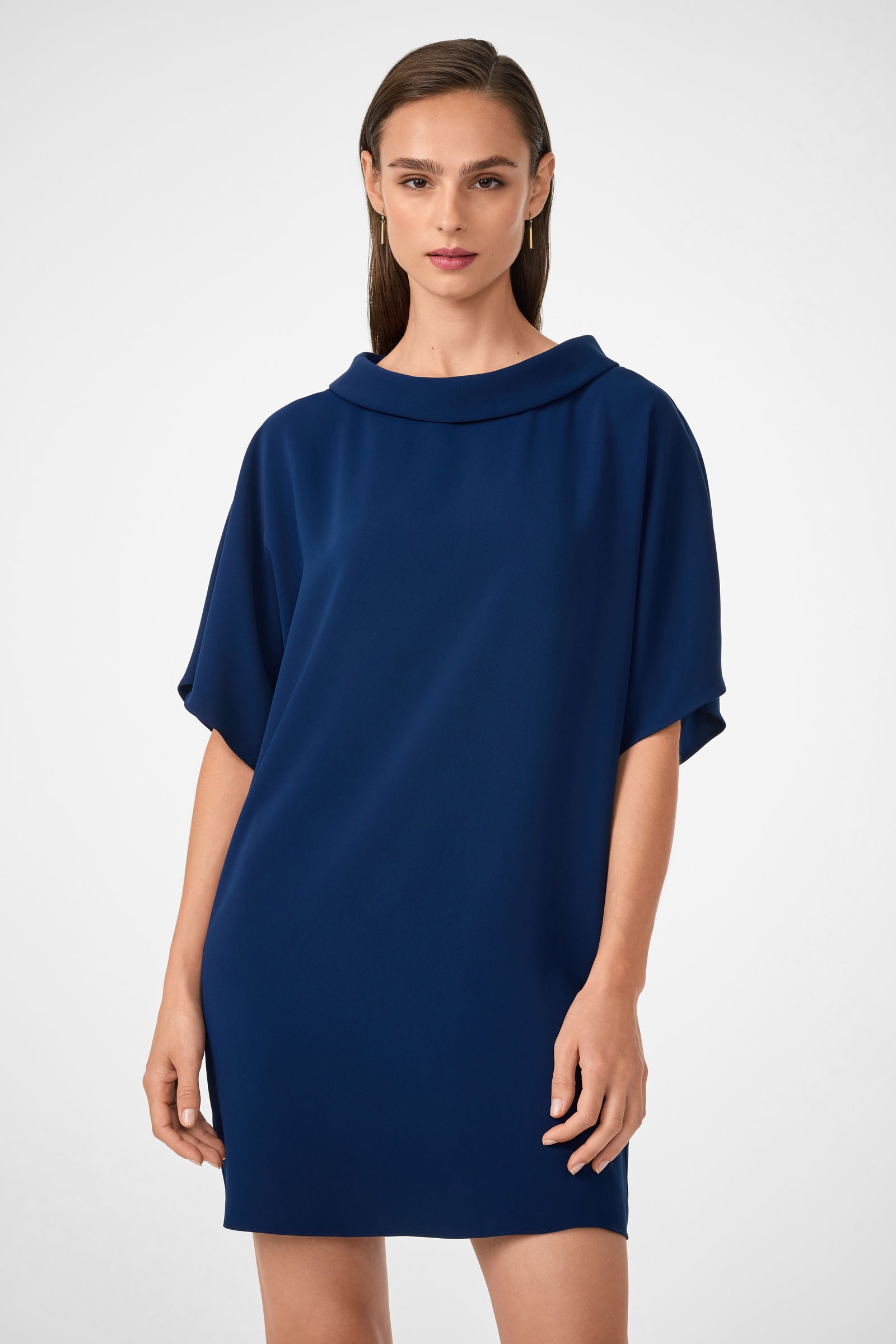 A woman with straight brown hair models the Honeysuckle Short Dolmain Sleeve Shift Dress, a loose-fitting, dark blue dress with short sleeves and a wide neckline, as she stands confidently against a plain white background.