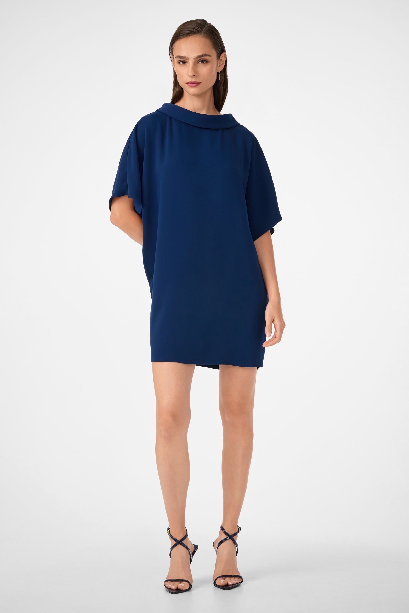 A woman stands against a plain background wearing the Honeysuckle Short Dolmain Sleeve Shift Dress in Ink, featuring a loose fit, short sleeves, and wide neckline. She pairs it with black strappy high-heeled sandals.