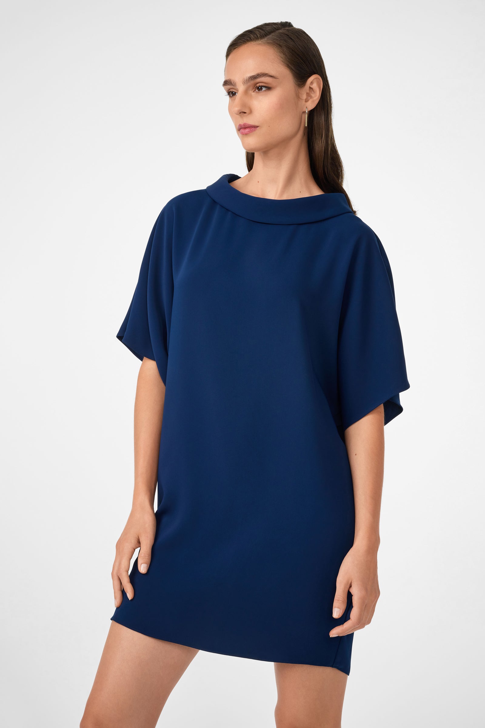 A woman wearing the Honeysuckle Short Dolmain Sleeve Shift Dress, a loose-fitting dark blue piece with short sleeves and a wide collar, stands against a plain light gray background, gazing slightly to the side.