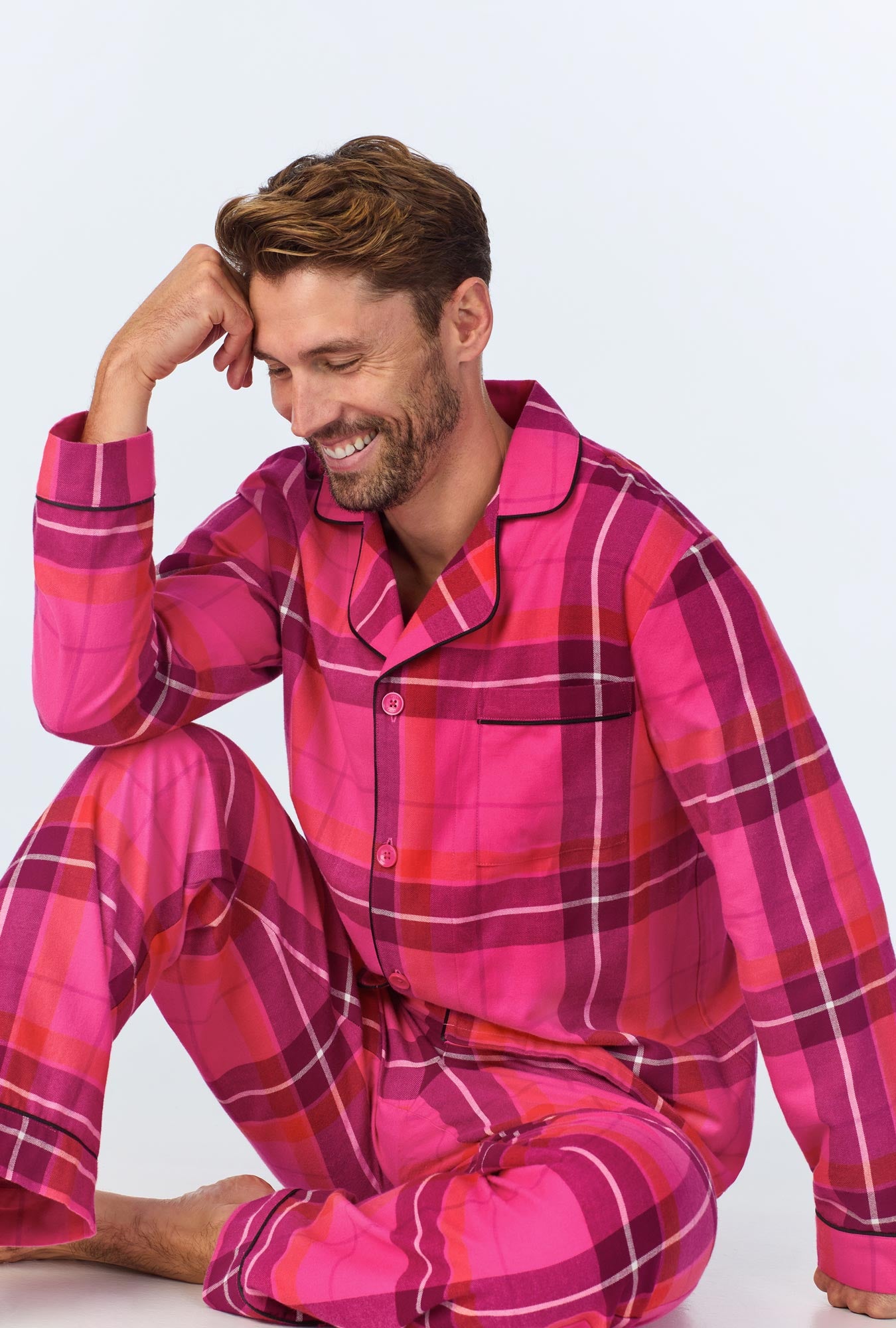 A man with short brown hair and a beard sits on the floor, smiling and resting his head on his hand. He is wearing the Aurora Plaid Mens Long Sleeve Long Pant Flannel PJ Set in Multi—a cozy choice for men’s sleepwear.