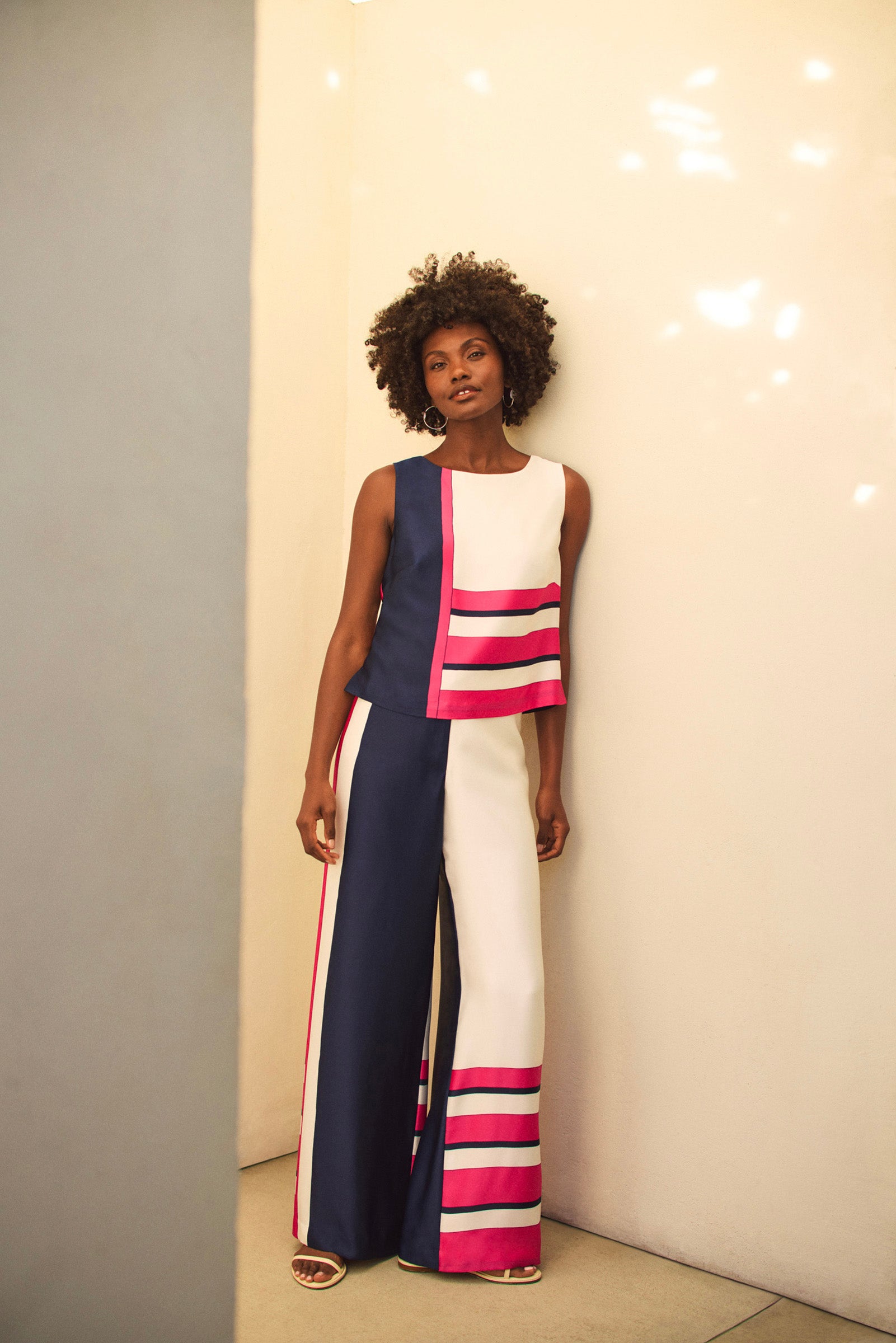 A woman with short curly hair stands against a light wall, wearing the Cadorna Linee Sleeveless Top and wide-leg pants with navy, white, and bright pink color blocks and stripes. She completes the look with sandals.