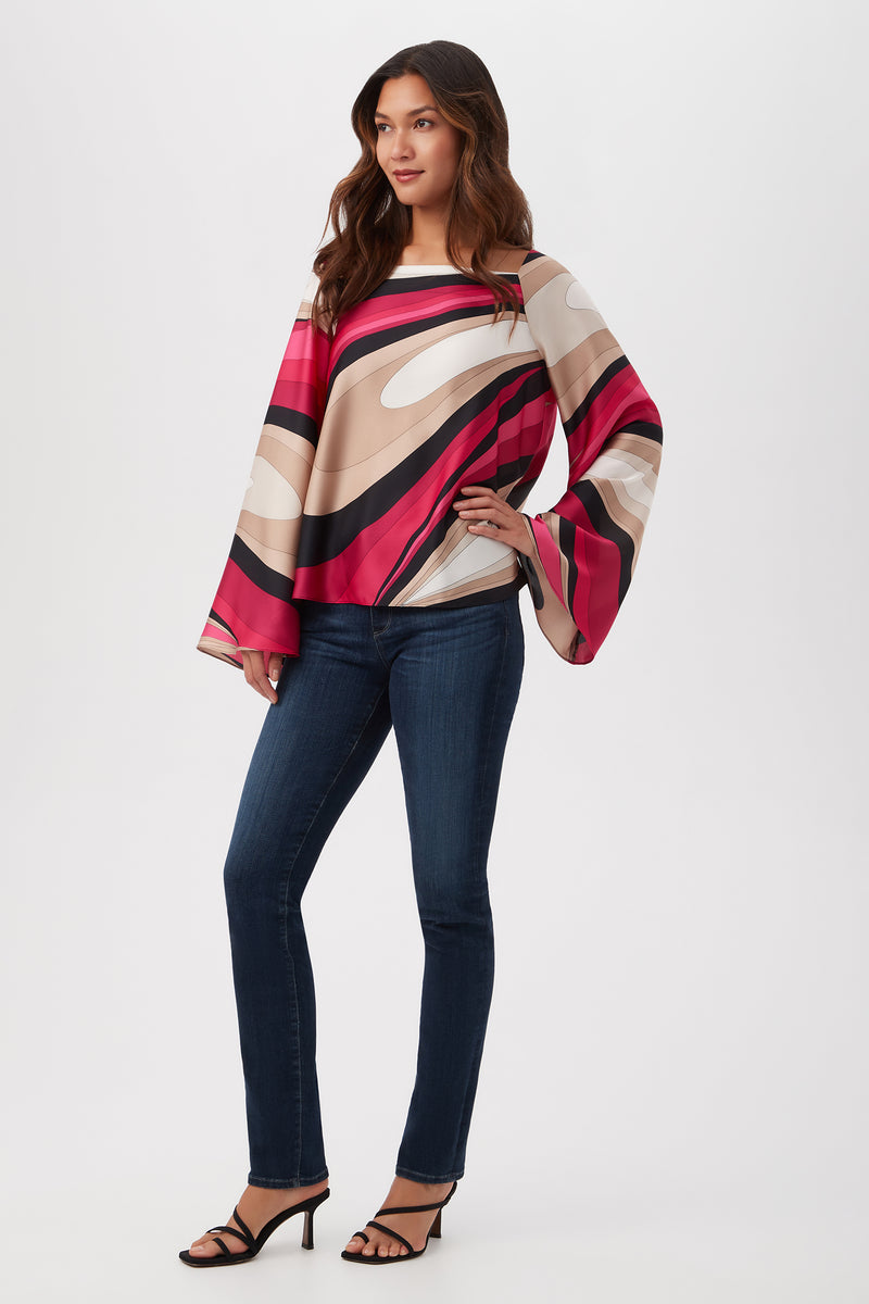 A woman wears the Torie Top - Wynwood Waves Satin Georgette, a long-sleeve, loose, multicolored top with bold red, pink, beige, and black swirls, styled with dark blue jeans and black heeled sandals against a white background.