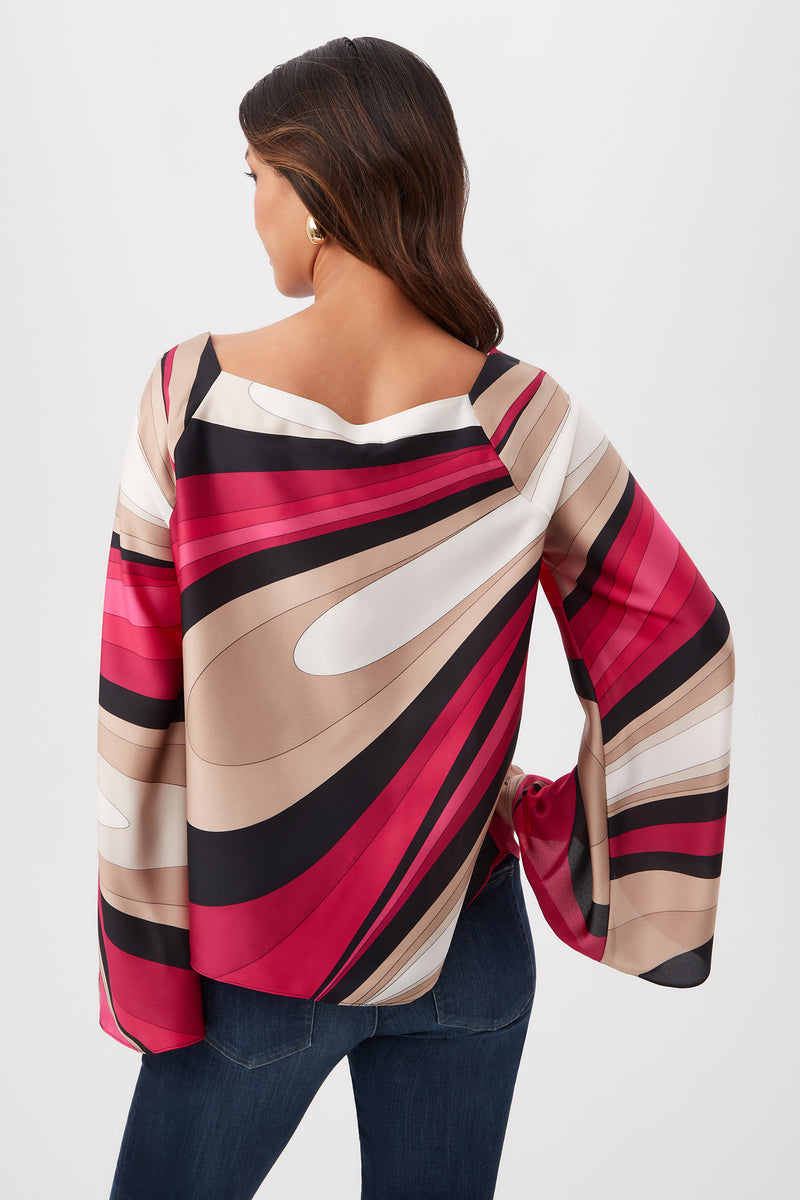 A woman wearing the Torie Top - Wynwood Waves Satin Georgette with abstract red, black, beige, and white patterns is facing away to show the back of the blouse and her wavy dark hair. She pairs it with blue jeans.